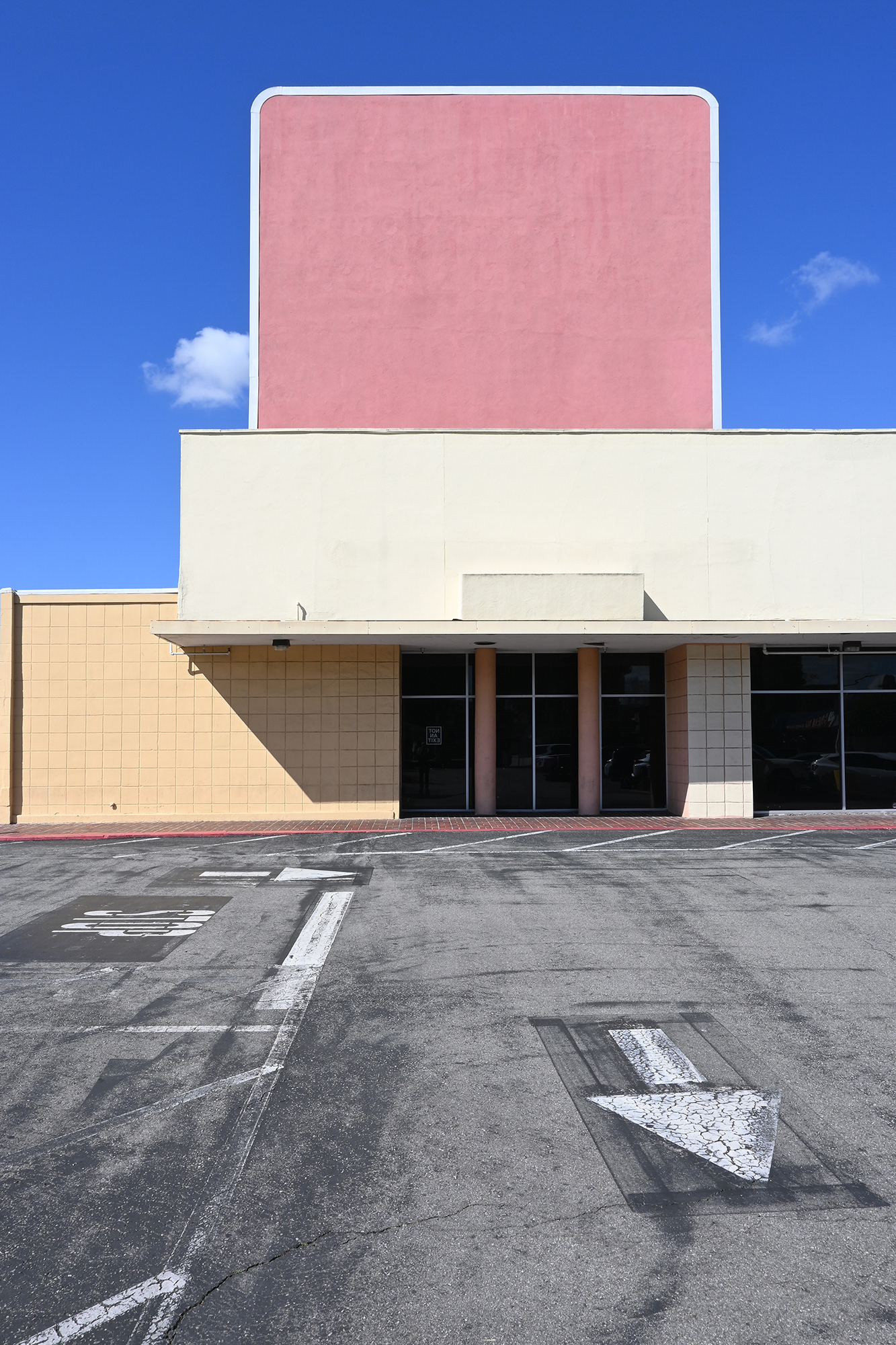 Photograph of a closed department store in El Camino Plaza, Old Town Tustin. The buildign has a large pink sign agaist a blue sky with small clouds the foreground is ashphalt with white lines and arrow.