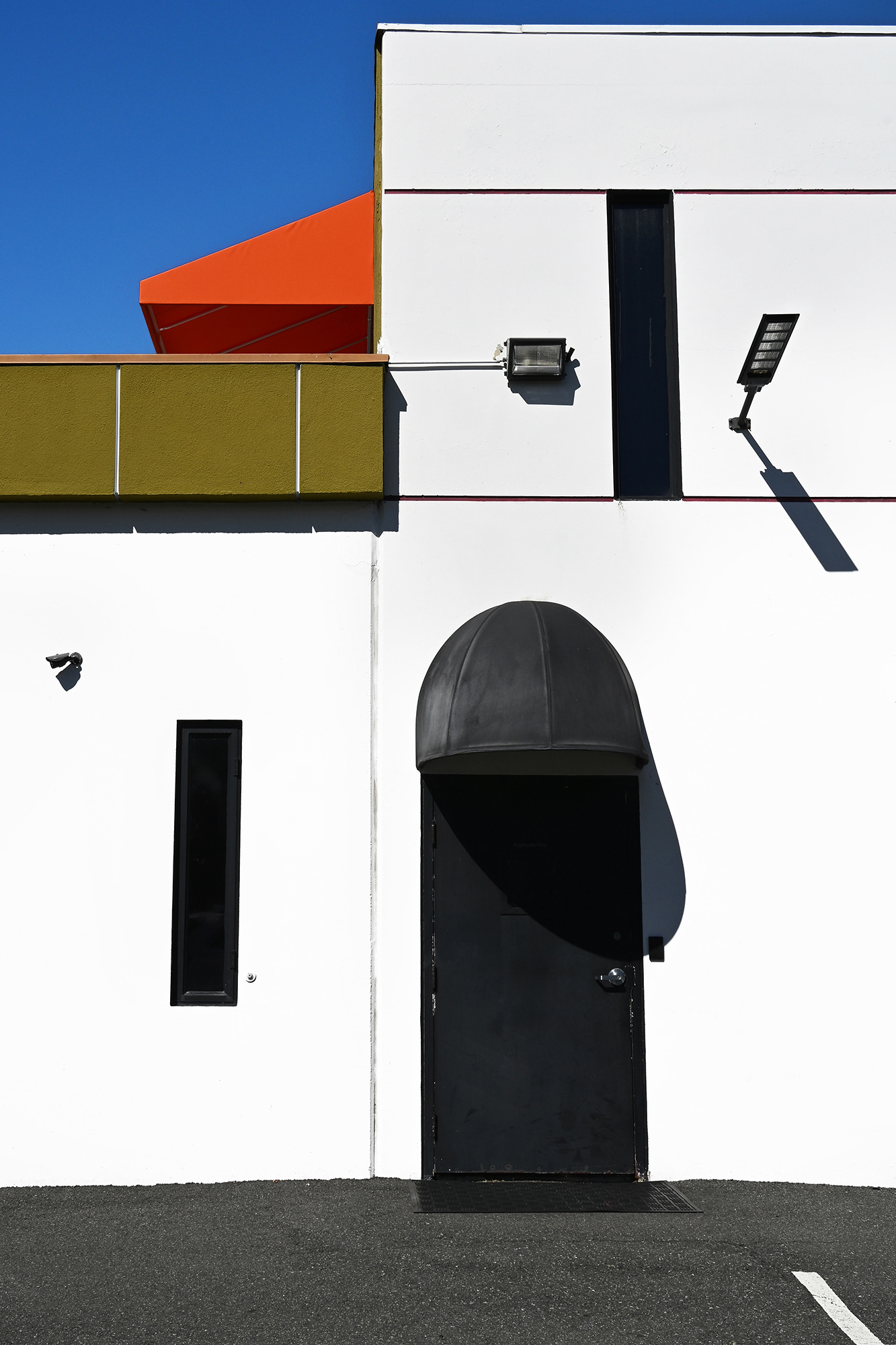 Photograph of a Black Door and Awning on a white building, above is another orange awning and green overhang, Irvine industrial Park