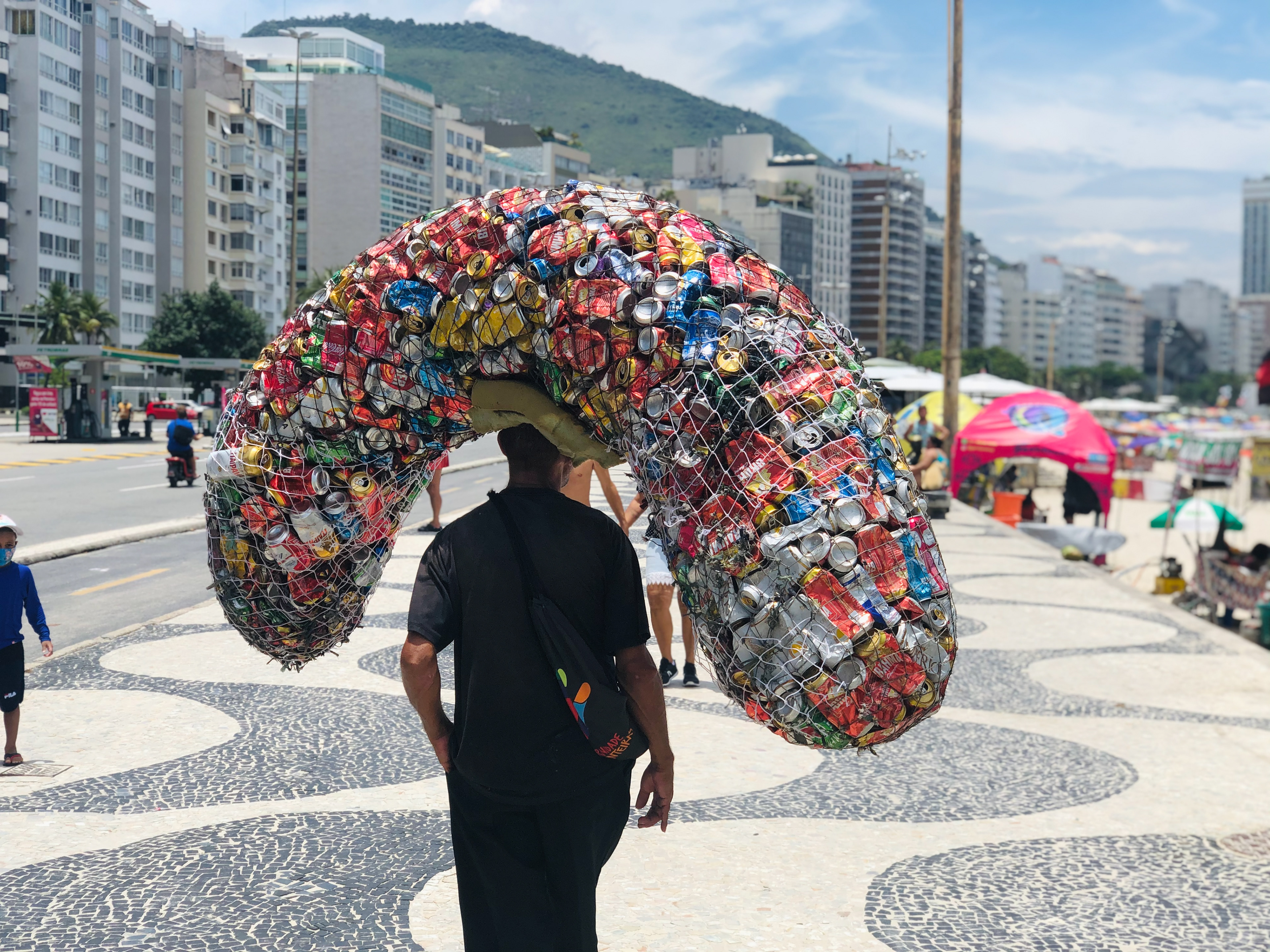 A man walks on Copacabana promenade with a large sack of aluminum cans on his head.