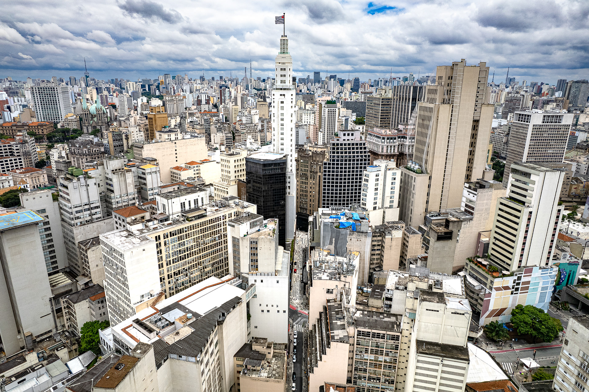 Aerial view of central São Paulo dominated by the historic Altino Arantes building, once the tallest in Latin America, rising among a dense urban skyline under dramatic clouds.
