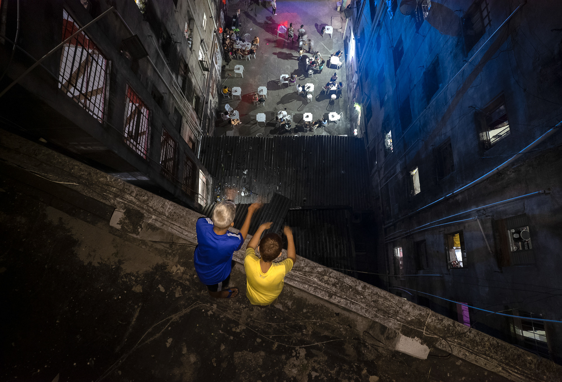 Two boys sitting on the edge of a rooftop inside the Occupy Mauá building, looking down at a courtyard filled with people during a night event in central São Paulo.