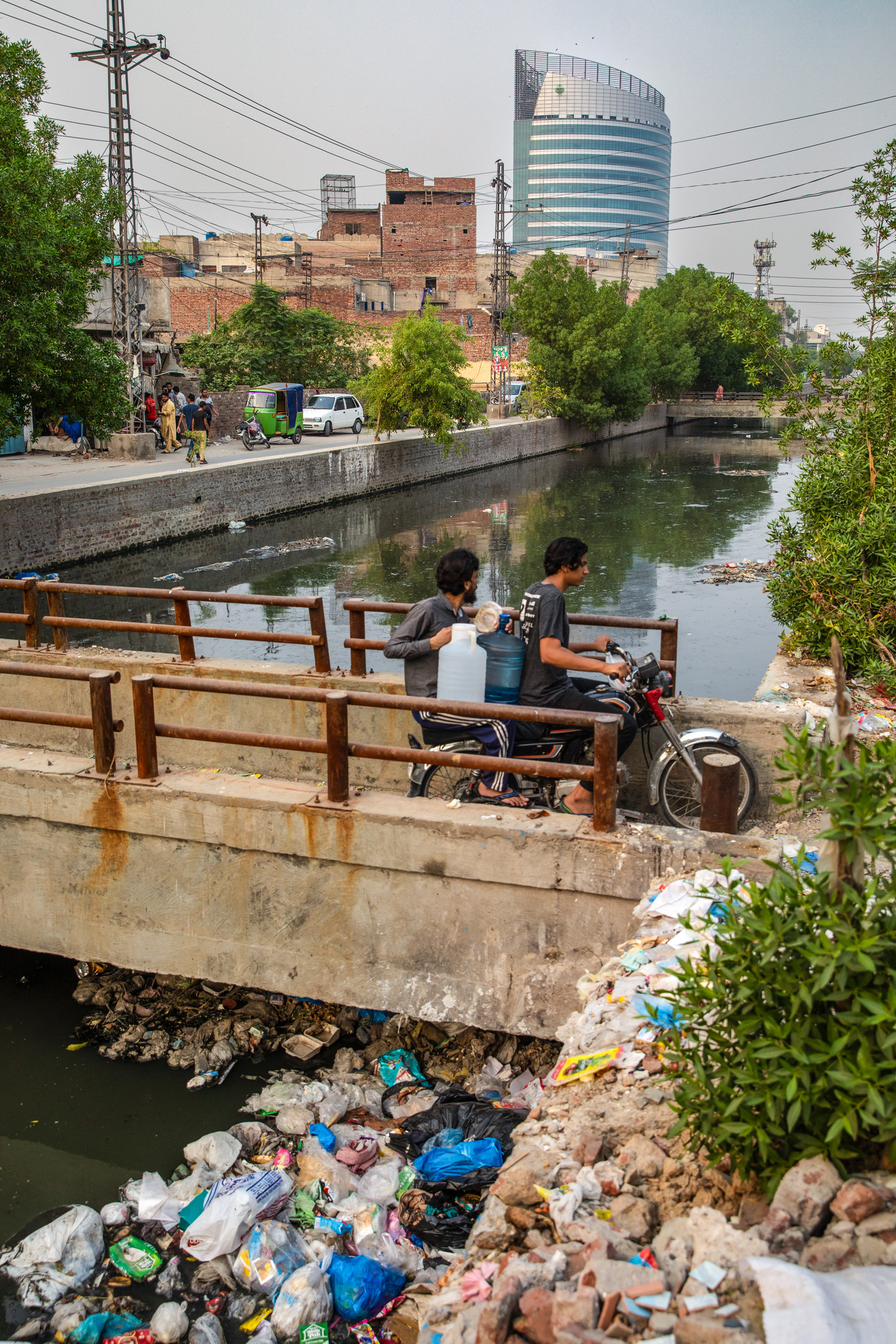 Two men on a motorbike going over a bridge on a river full of trash.