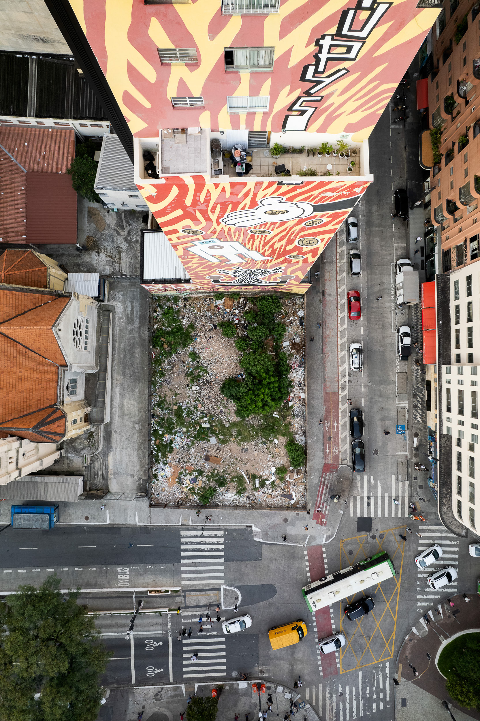 Aerial view of a vibrant mural on a high-rise in central São Paulo, overlooking a fenced-in lot filled with debris, highlighting the contrast between art and urban neglect.