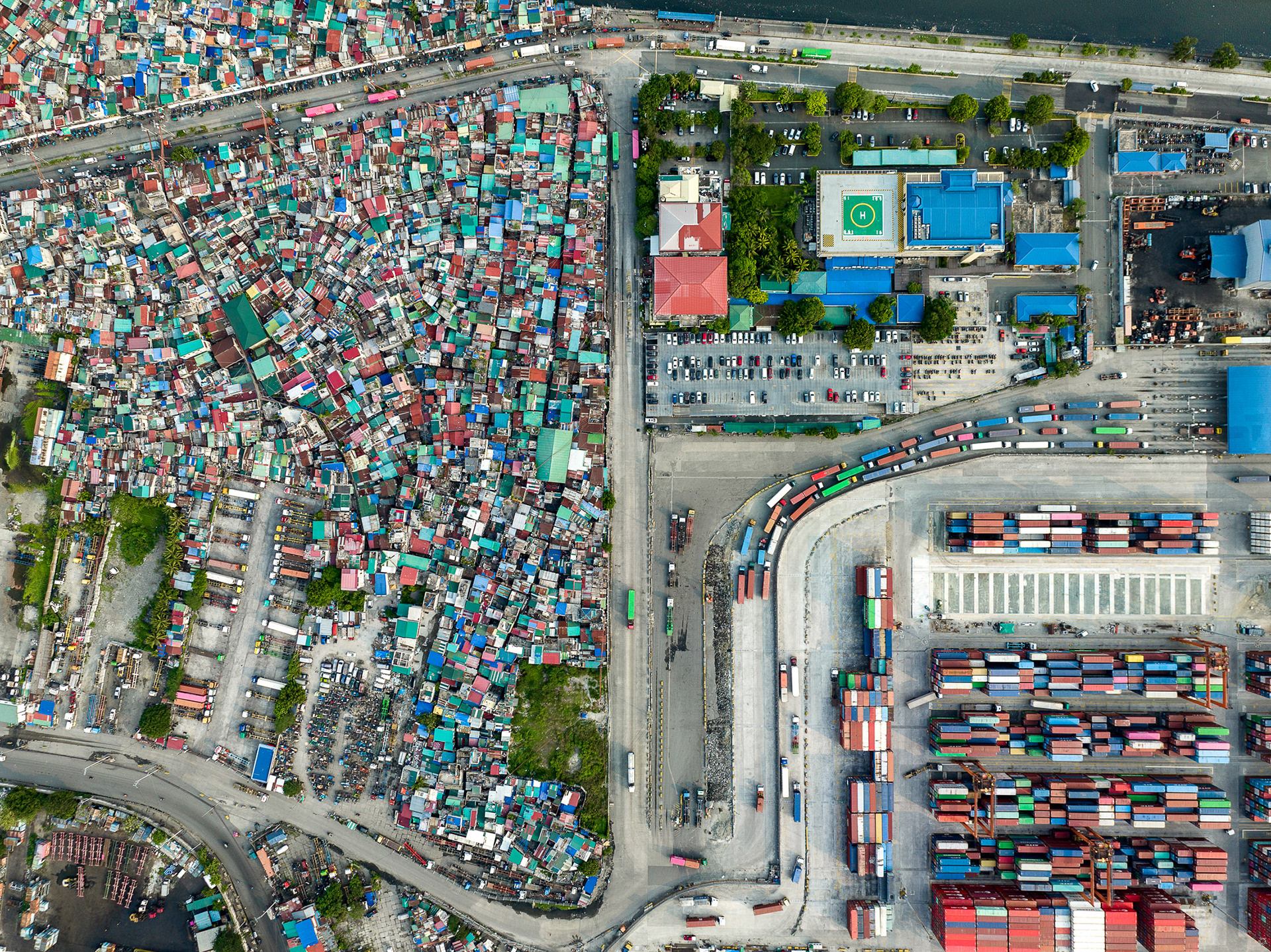 Top-down drone image of Manila showing a tightly packed informal neighborhood adjacent to a major shipping yard, bisected by a road filled with truck traffic entering the port.