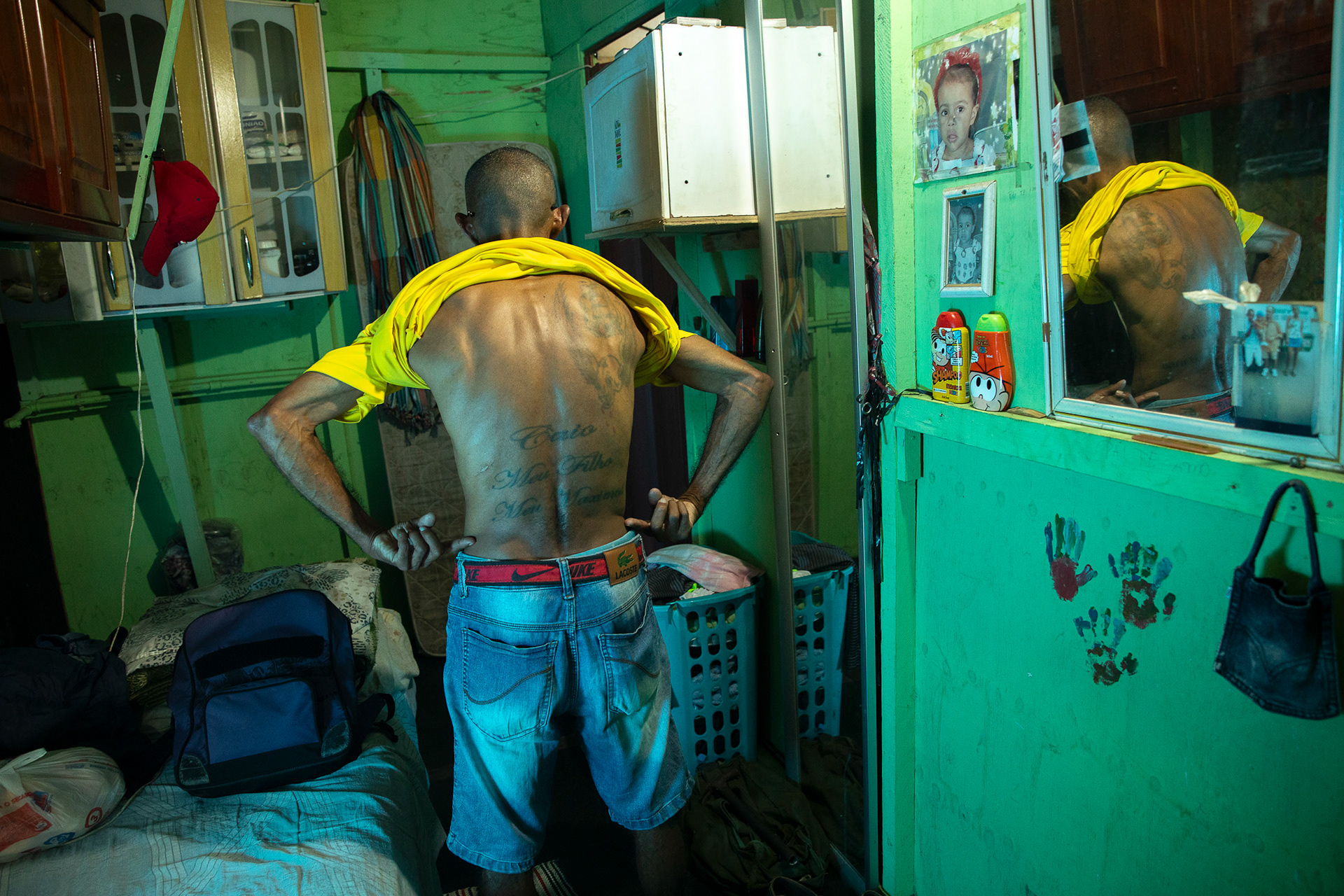 Inside a stilt house in Santos, a man prepares for the day in a cramped room, his back tattooed with his son’s name, a symbol of survival and love.