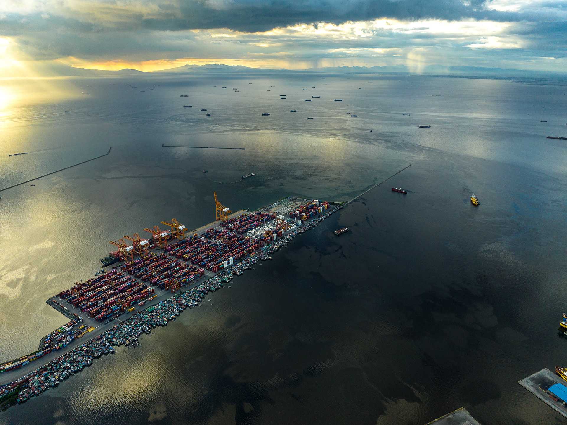 Wide-angle drone view of the Port of Manila at sunset, with container terminals and shipping lanes surrounded by calm water, highlighting nearby informal dwellings on port-controlled land.