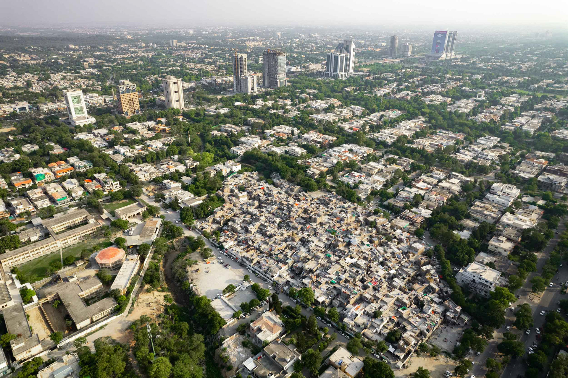 A drone view of a slum inside a nice forested neighborhood next to skyscrapers.