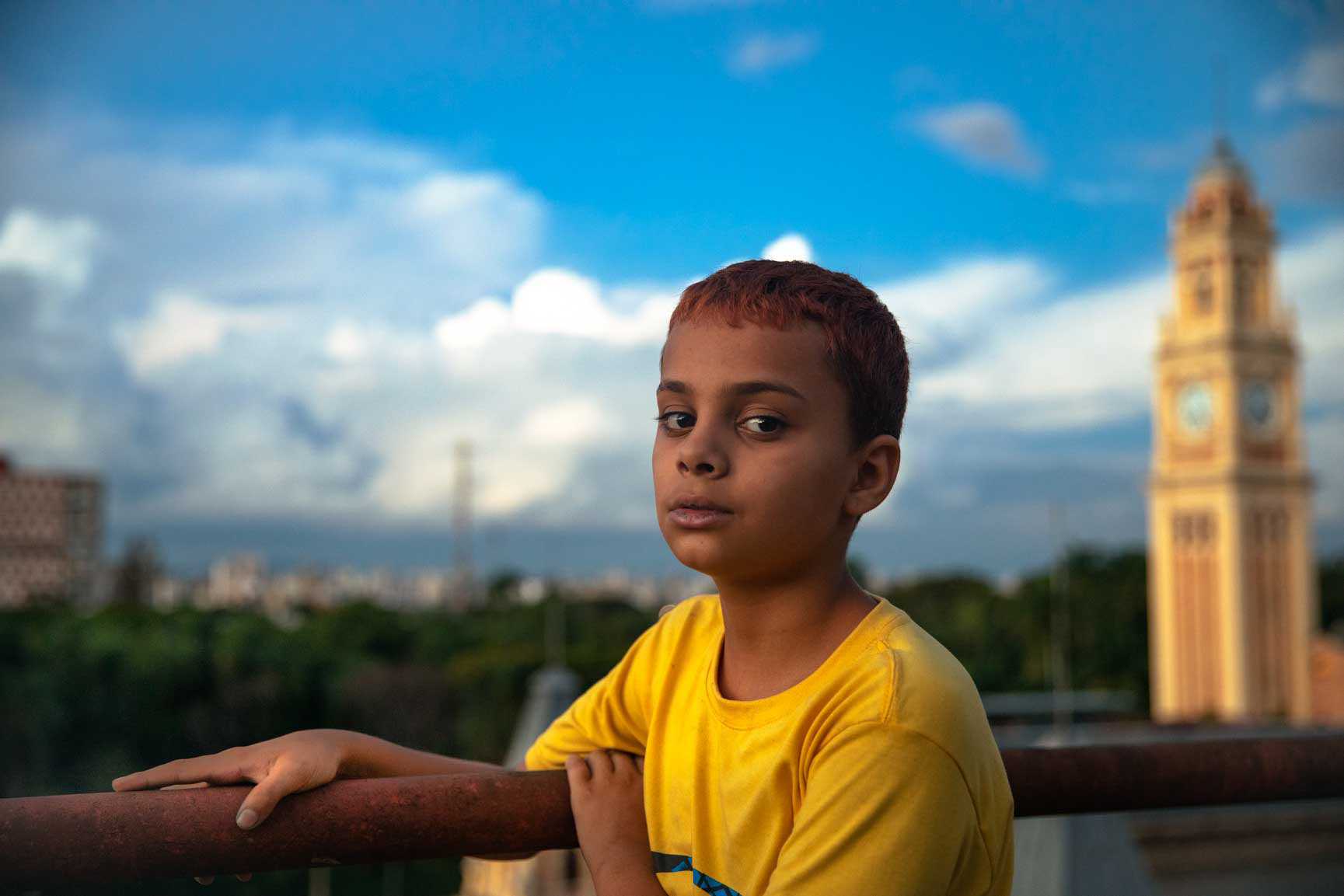 Portrait of a young boy named Gilmar standing on a balcony at Ocupação Mauá, with São Paulo’s Luz train station clock tower in the background.