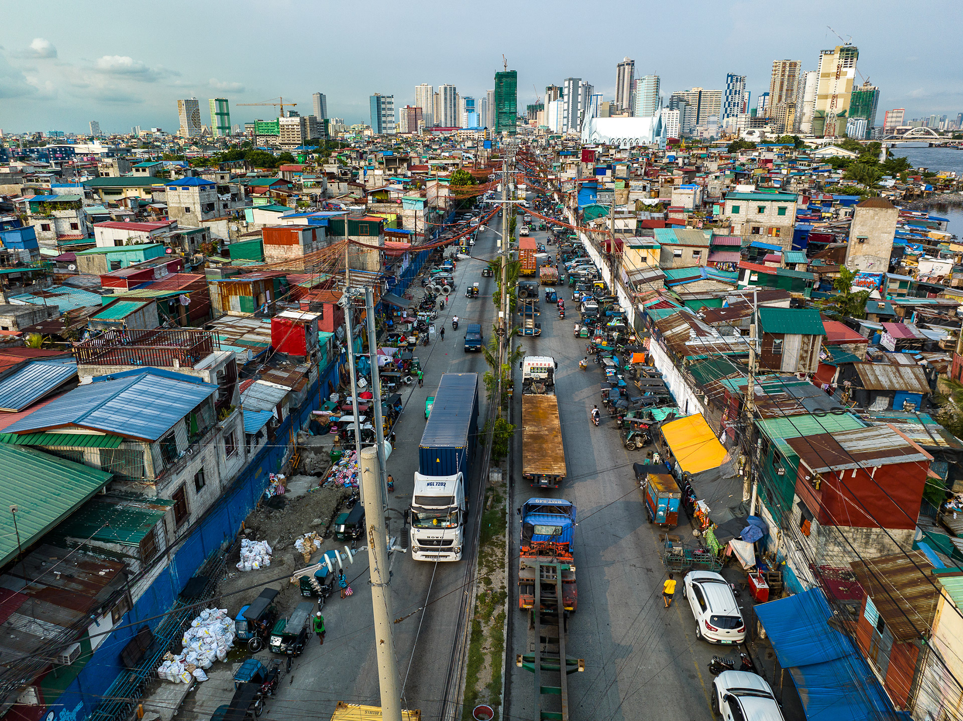 A busy road cutting through informal housing in Manila, crowded with trucks and power lines, leading toward the high-rise skyline in the background.