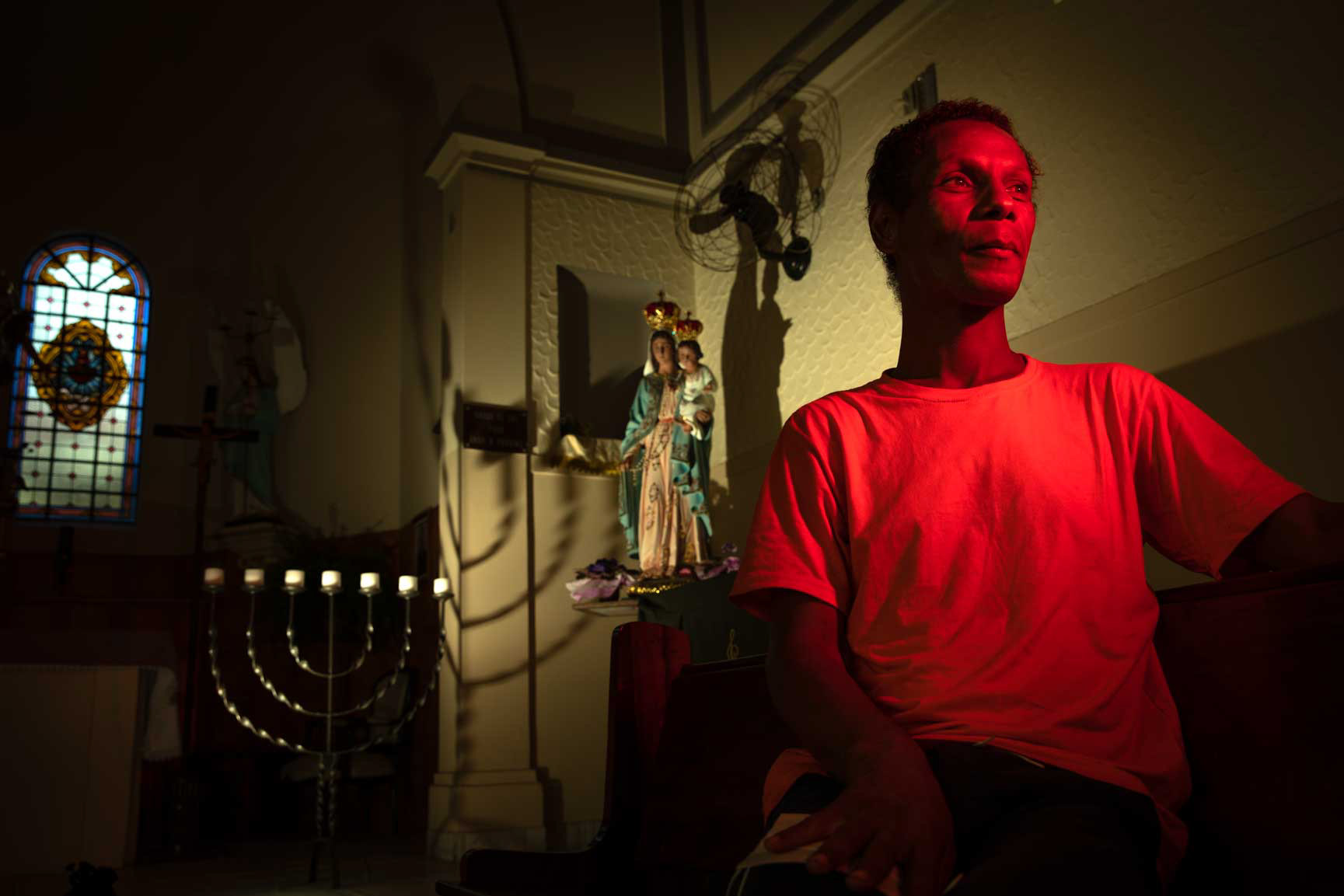 Portrait of Alessandra Martinelli, a trans woman living with HIV, sitting inside a dimly lit room in Mooca, São Paulo, with soft red light casting shadows on the wall behind her and a religious statue in the background.