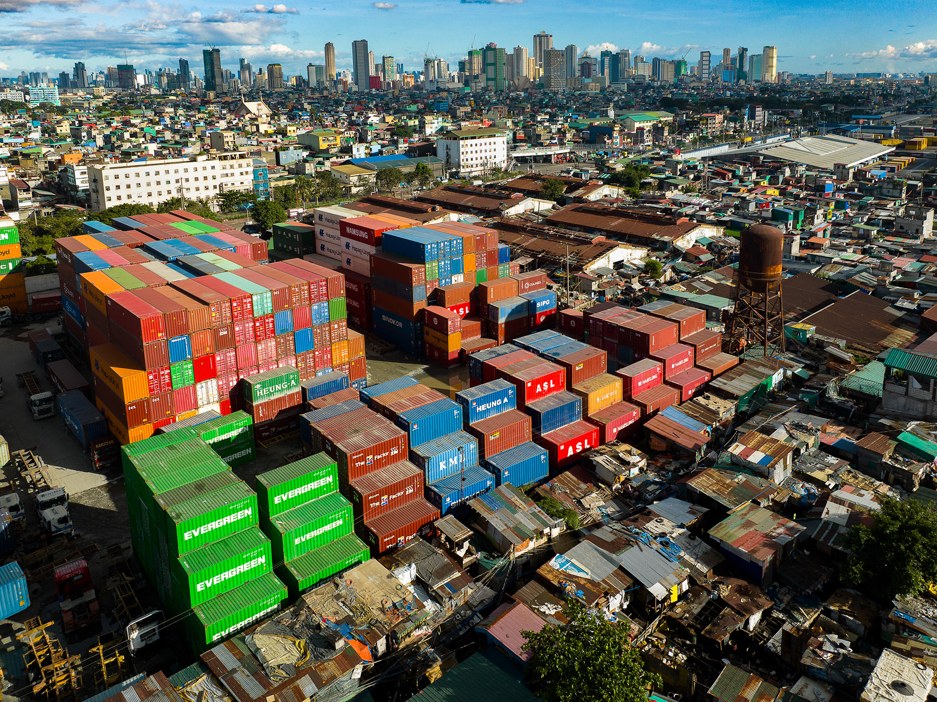 Overhead view of colorful shipping containers stacked beside an informal settlement at the Port of Manila, with Manila's skyline rising in the background.