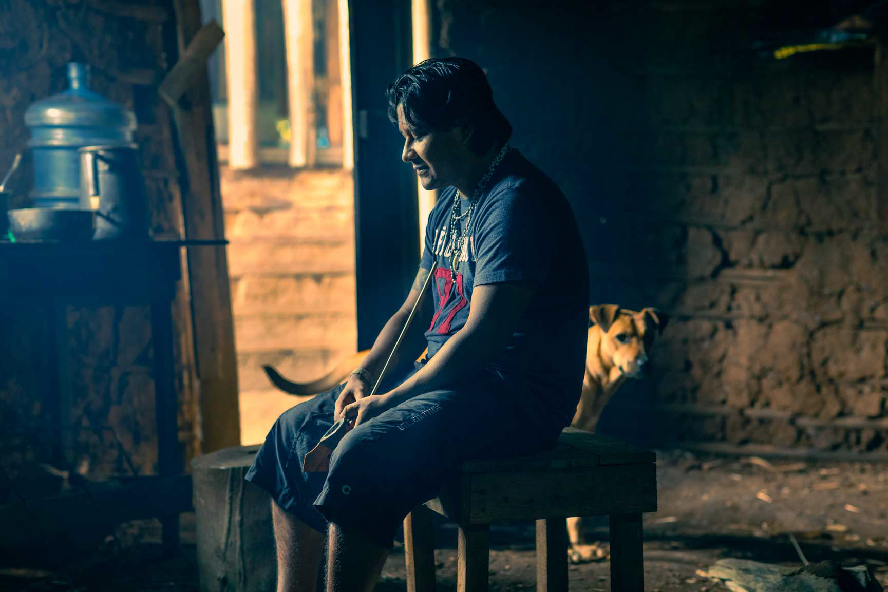 A Guarani man named Davi sits solemnly in a dimly lit room, representing the intersection of Indigenous identity, urban displacement, and mental health in São Paulo.