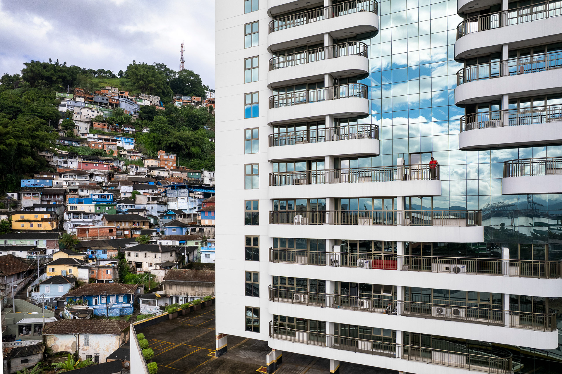 A stark visual of inequality as a favela clings to a forested hill while a mirrored high-rise reflects blue skies and private balconies in Santos.