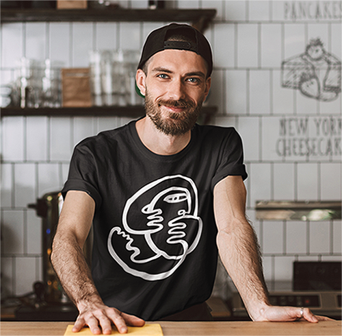 Young man in a baseball cap wearing a black T-shirt with a white minimalist abstract face artwork while standing behind a café counter.