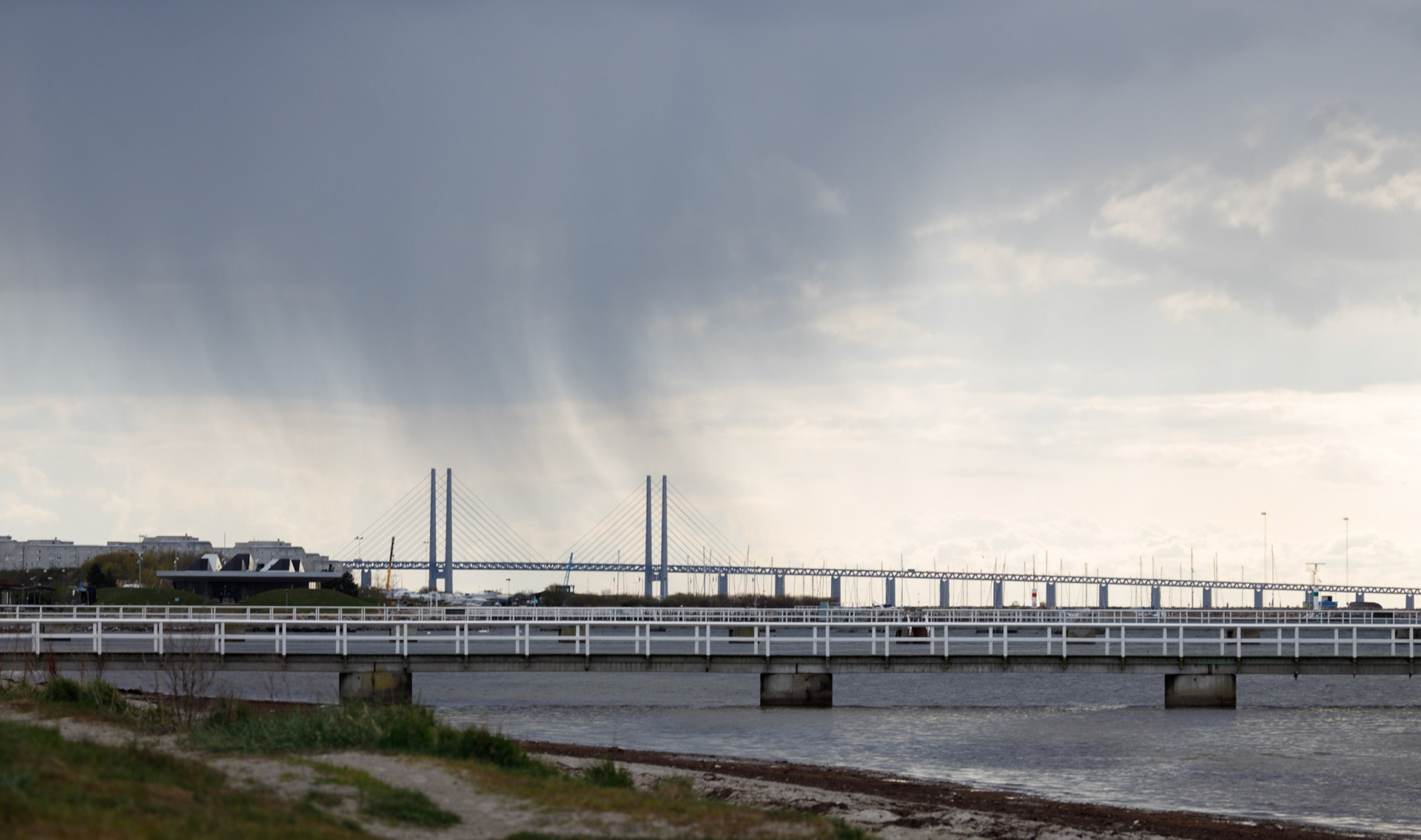 Ribersborgstranden, Ribban, brygga 4 även kallad Kaffebryggan. Bryggorna på Ribban är numrerade 1 till 10, medan bryggorna på Sibbarp är bokstaverade från A till N. Praktiskt för räddningstjänsten som inte behöver fundera på vilken strand de ska köra till. I bakgrunden syn ett regnväder över Oresundsbron. Mellan bryggan och bron till vänster syns Utställningsbyggnaden som användes för utställningar när bron byggdes.