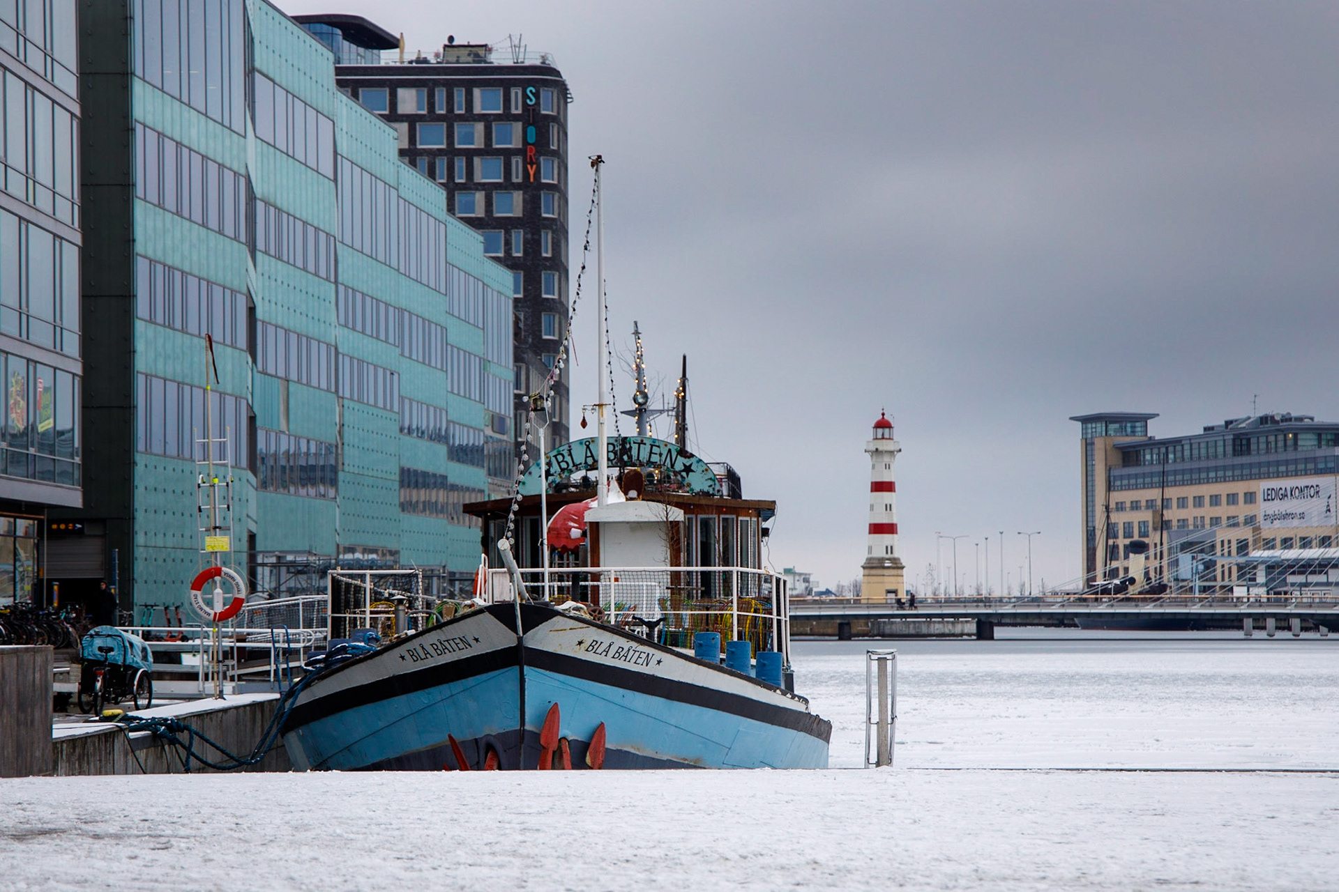 Blå båten är byggd 1888 i Holland där hon transporterade gods på Västeuropas floder. 1997 kom hon till Malmö och har sedan dess använt som musikställe. Hon finns längst inne i Inre hamnen. I bakgrunden syns Malmö Inre fyr som byggdes 1878 och lyste fram till 1936. Till höger om fyren syns Smörkontrollen som byggdes 1934. Fram till 1991 kontrollerades svenskt smör här.