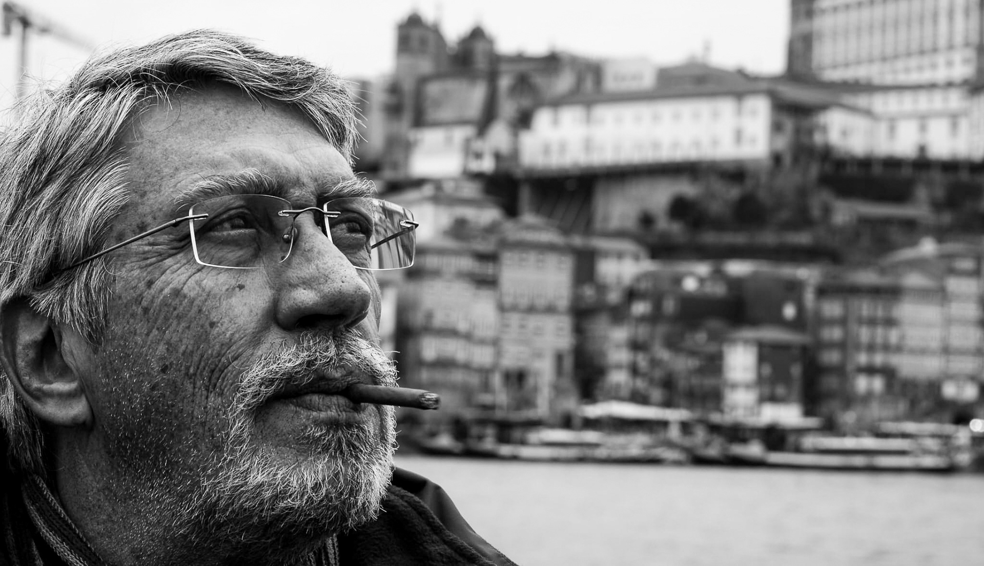 Black and white portrait of an older man with glasses smoking a cigar, looking into the distance with a blurred riverside city background.