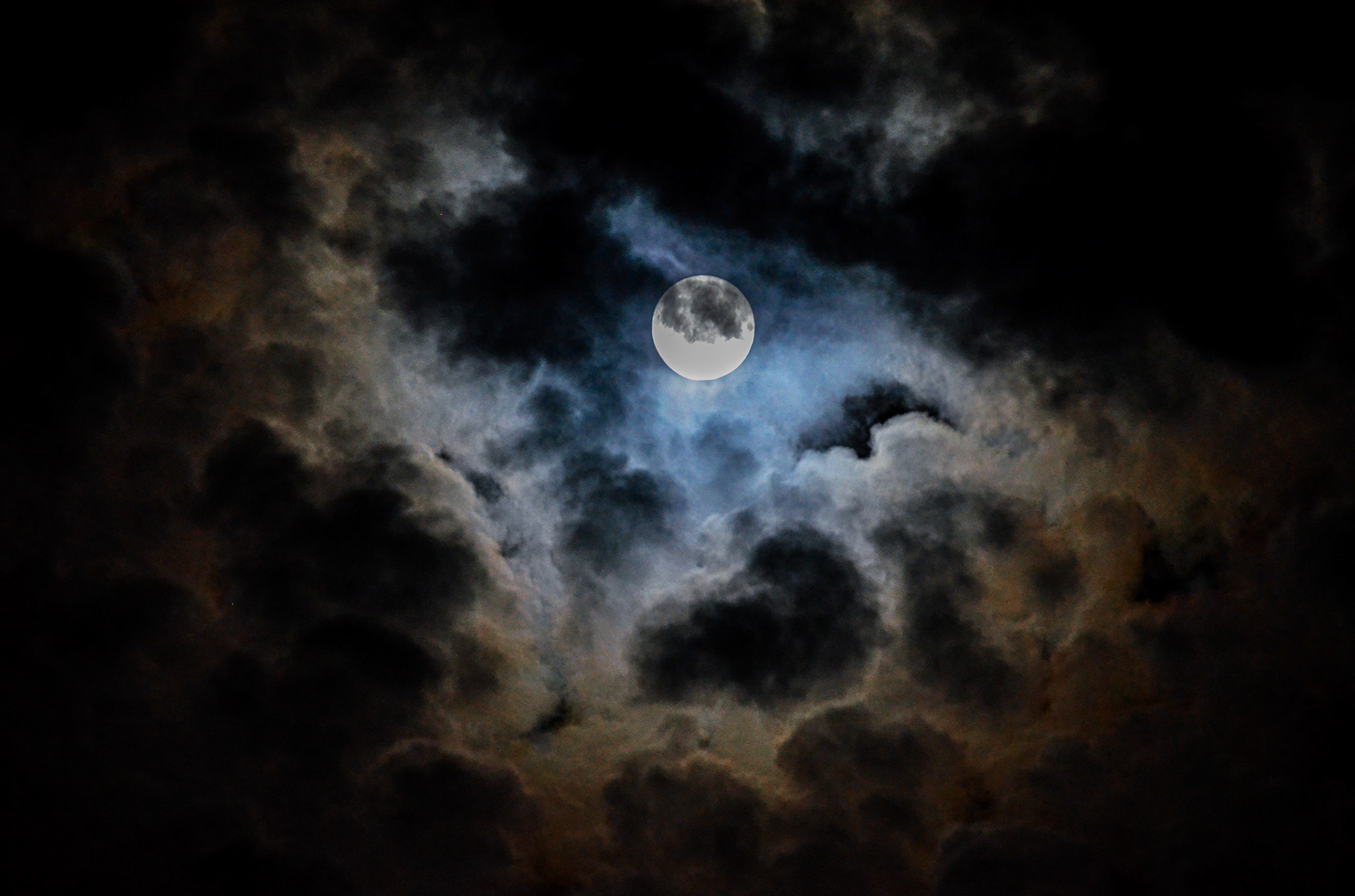 Full moon partially obscured by dramatic clouds, surrounded by cool blue and warm amber tones in a moody atmospheric night sky.