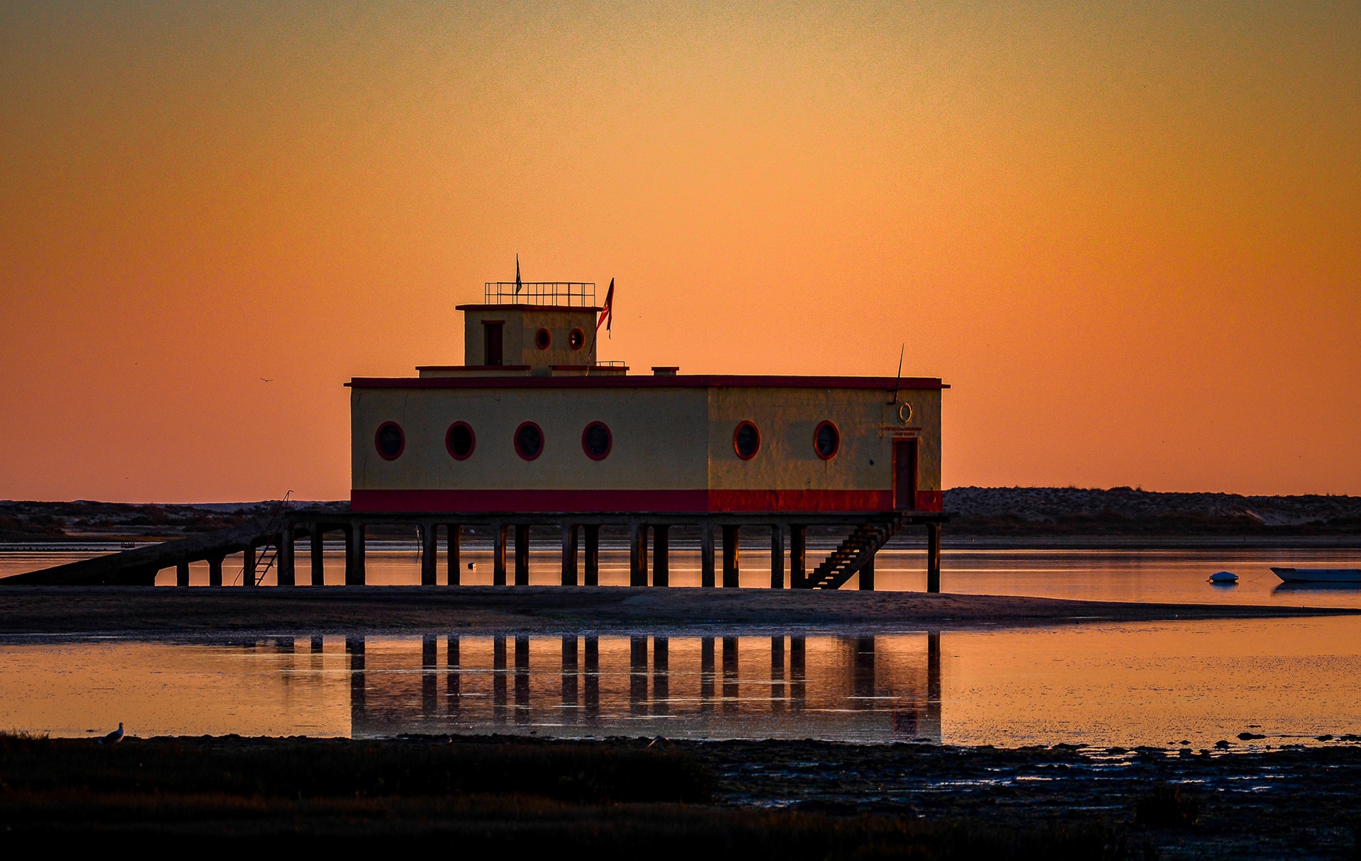 Minimal architectural structure standing over calm tidal waters at sunset in Fuseta, Algarve, reflected in still water beneath an orange sky.