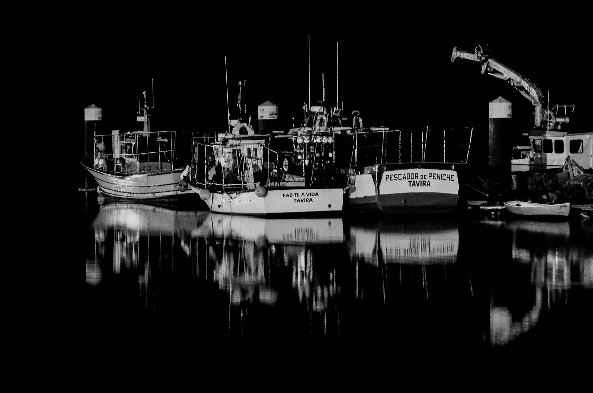 Black and white photograph of small boats moored at night with reflections in dark water