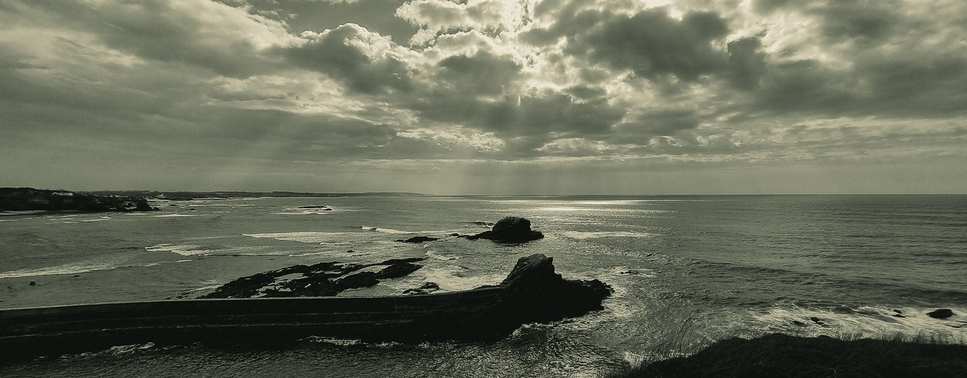 Rays of light emerging through heavy clouds above a quiet Atlantic coastline, creating an atmospheric monochrome seascape.
