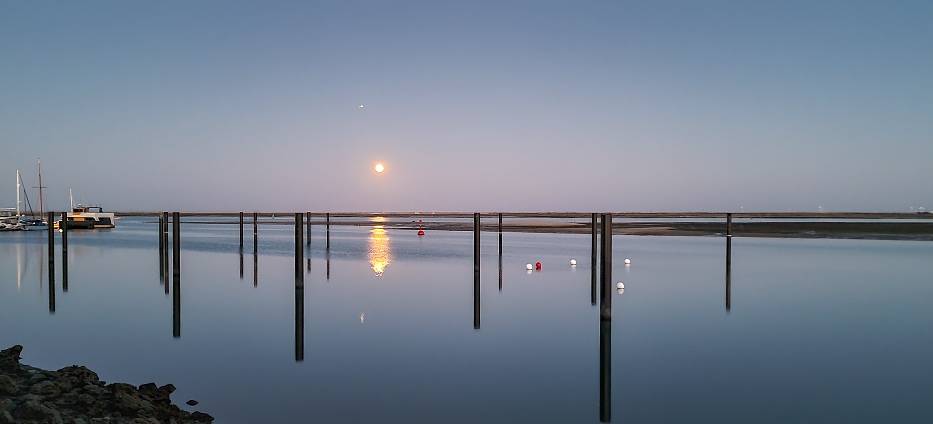 Full moon reflected on still water in Ria Formosa lagoon, with vertical wooden posts and a soft twilight sky.