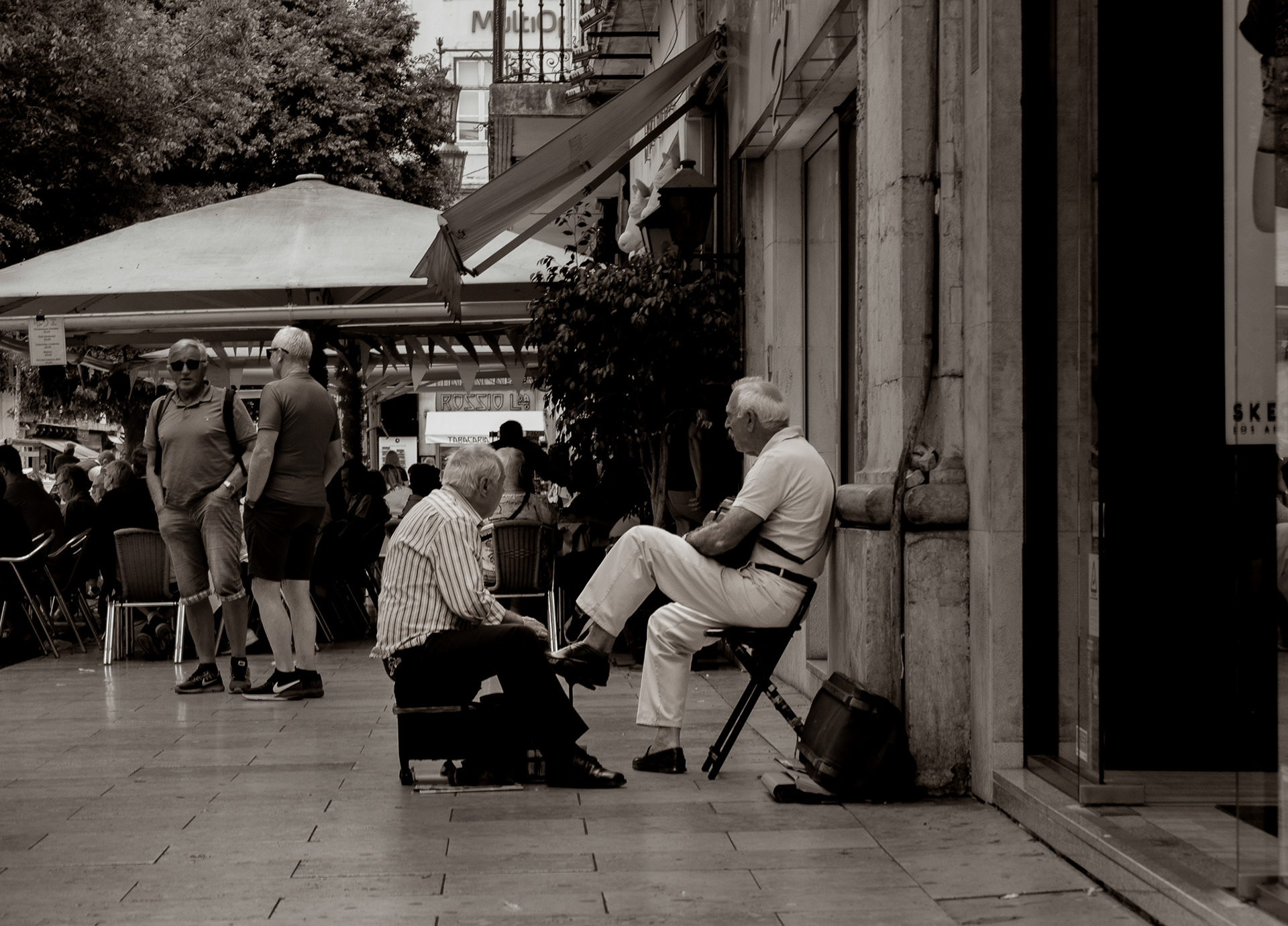 Two men in a quiet street interaction, one polishing the other's shoes, set against a busy urban background in Lisbon.