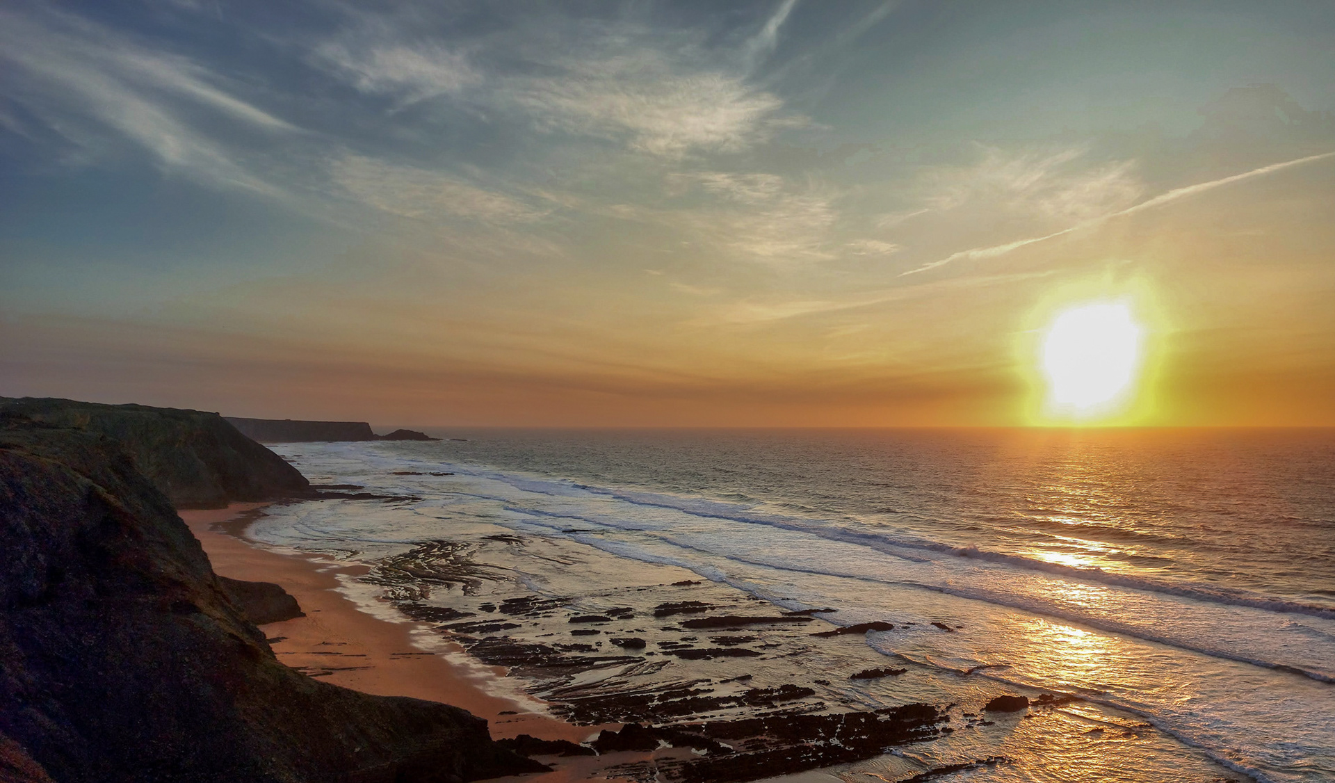 Sunset over Arrifana coast with cliffs, rocky shoreline and Atlantic waves reflecting warm golden light.