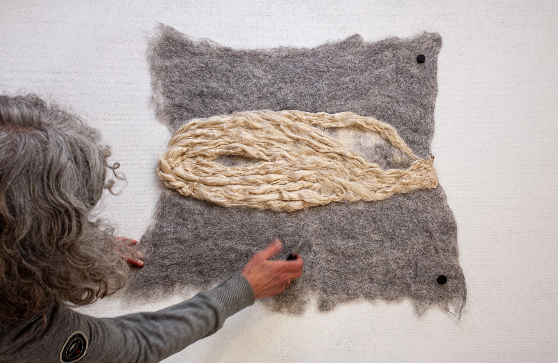 Artist adjusts a gray felt panel on the wall; an oval loop of golden Tussah silk is centered on the wool, secured with matte standoffs.