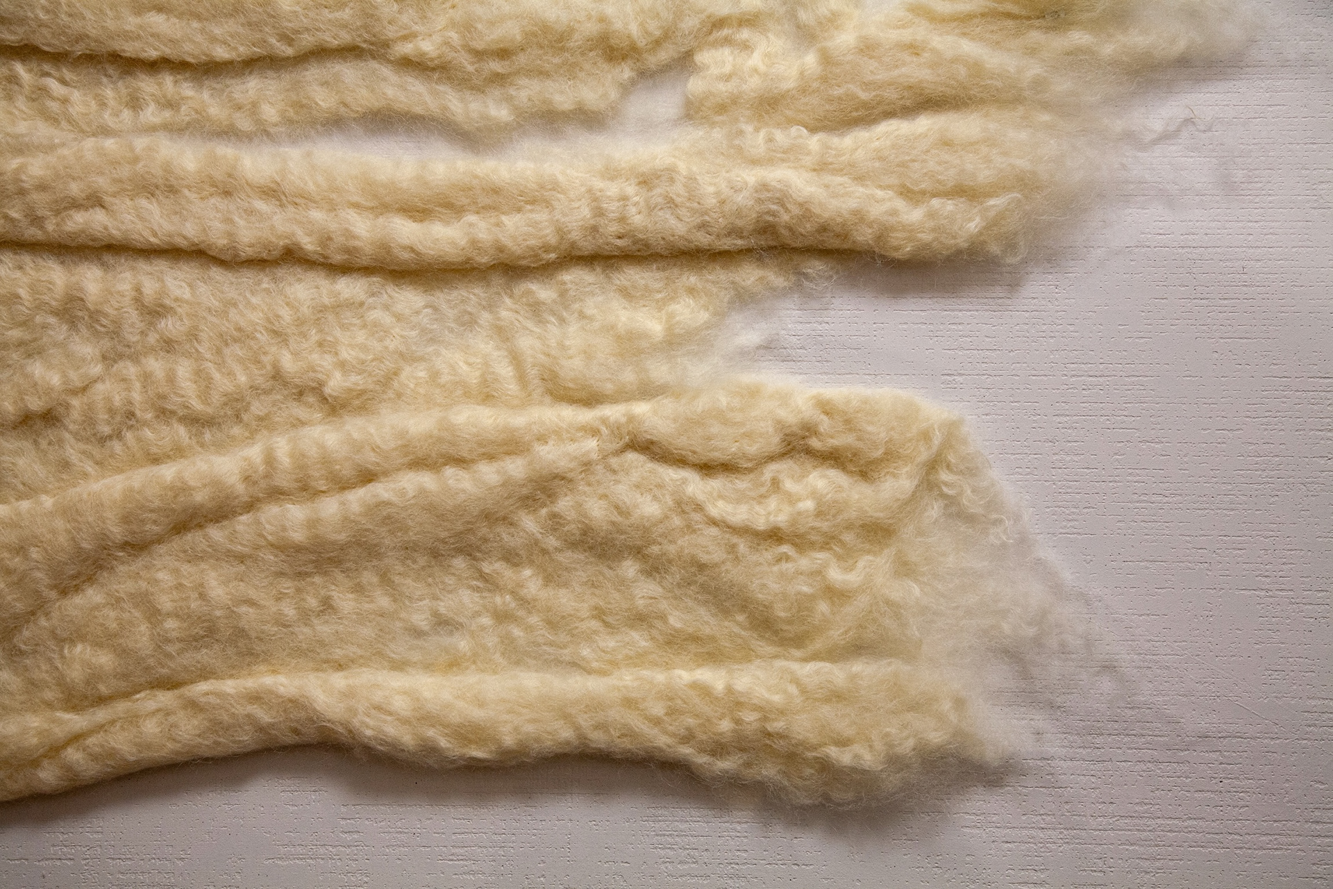 Close-up of layered cream felted wool bands with curly texture and wispy edges, resting on a clear mount with soft shadow on the white wall beneath.