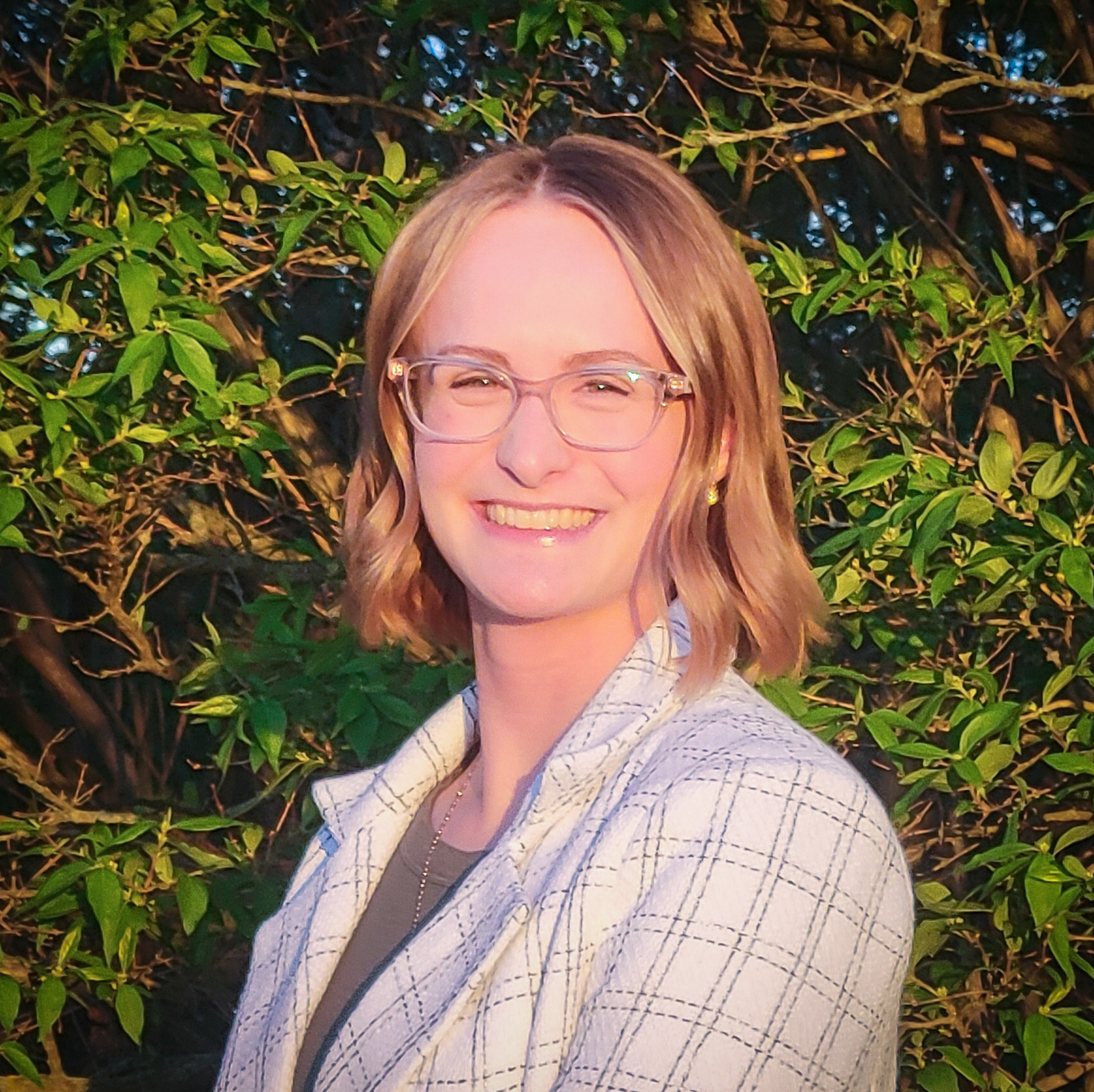 Head and shoulders portrait of young woman with short blonde hair and eyeglasses, wearing a white patterned blazer