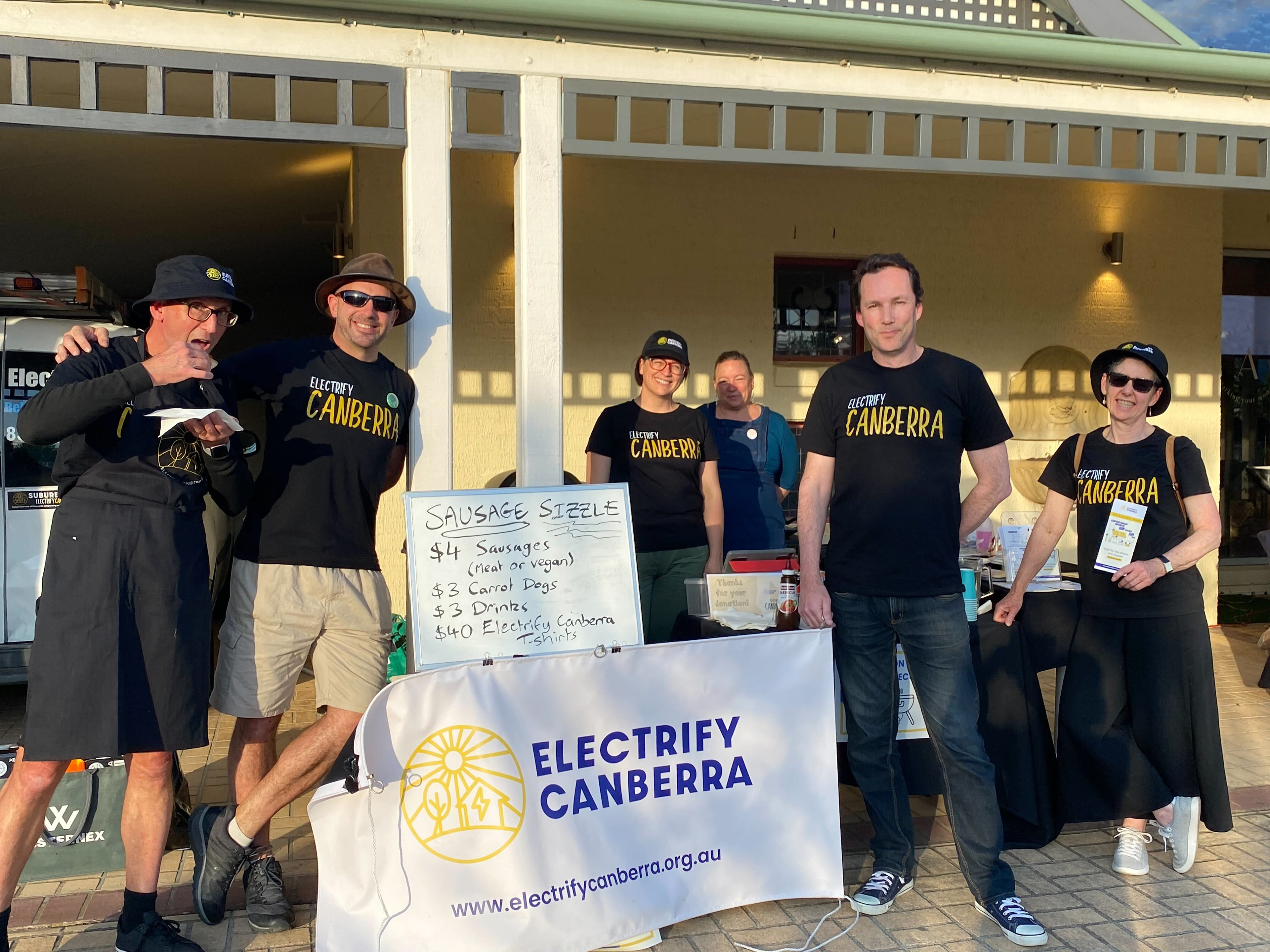 Image shows Electrify Canberra volunteers at a fundraising sausage sizzle with Electrify Canberra banner in the centre and each volunteer wearing their branded tshirt.