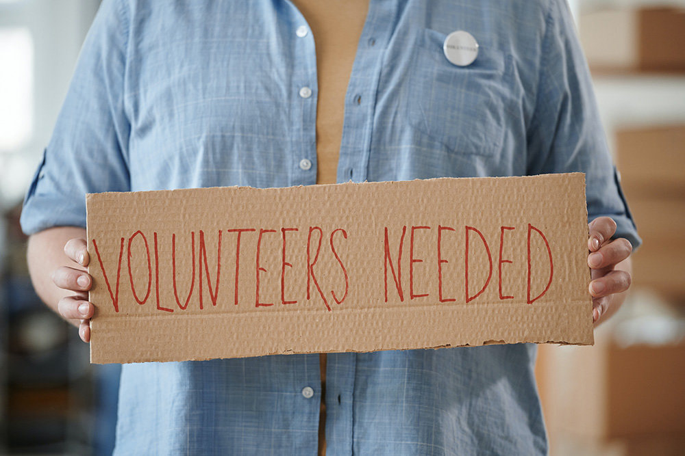 A person dressed in blue buttoned shirt holding cardboard with the words "volunteers needed" in red.