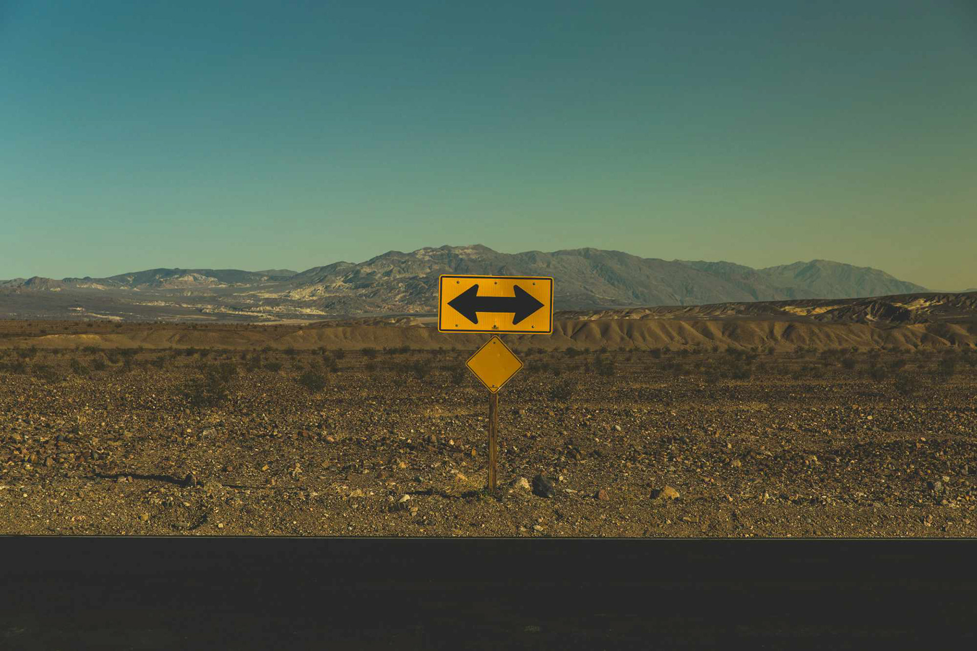 A yellow sign with arrows pointing opposite directions in a deserted location with a mountain in the background.