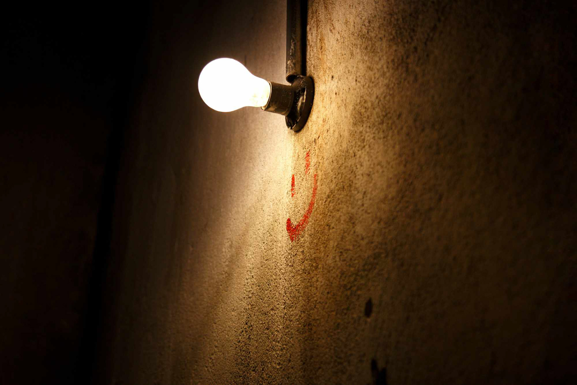 Lightbulb switched on highlighting a hand drawn smiley face in red on a grey wall.