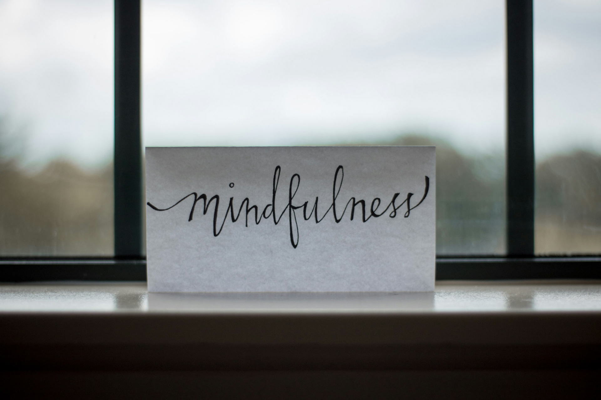 The word "mindfulness" handwritten in black on white paper positioned upright on a windowsill.