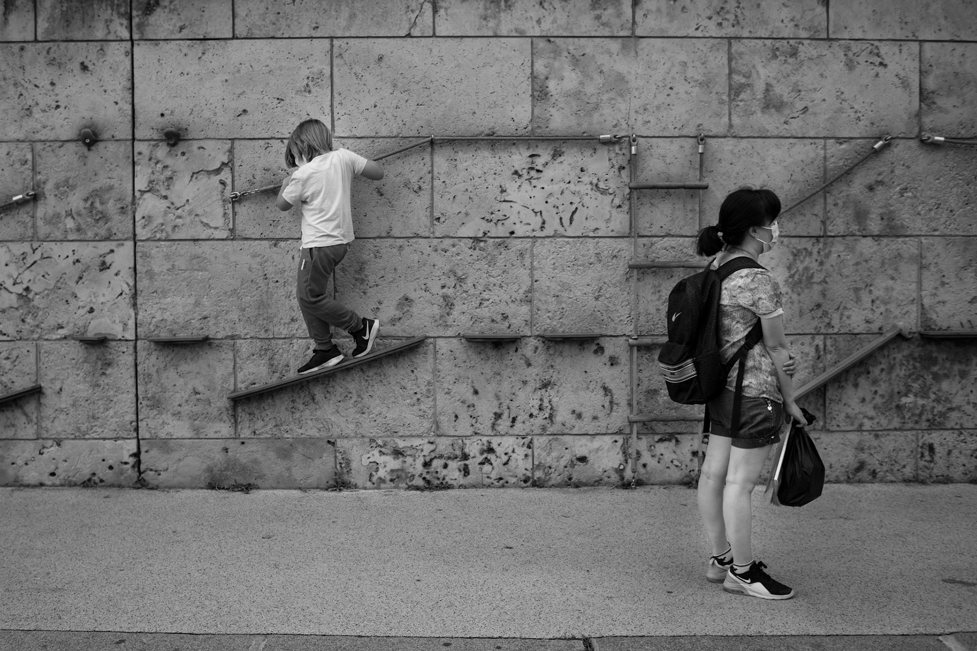 A boy playing at a public climbing wall while his mother waits absent-mindedly