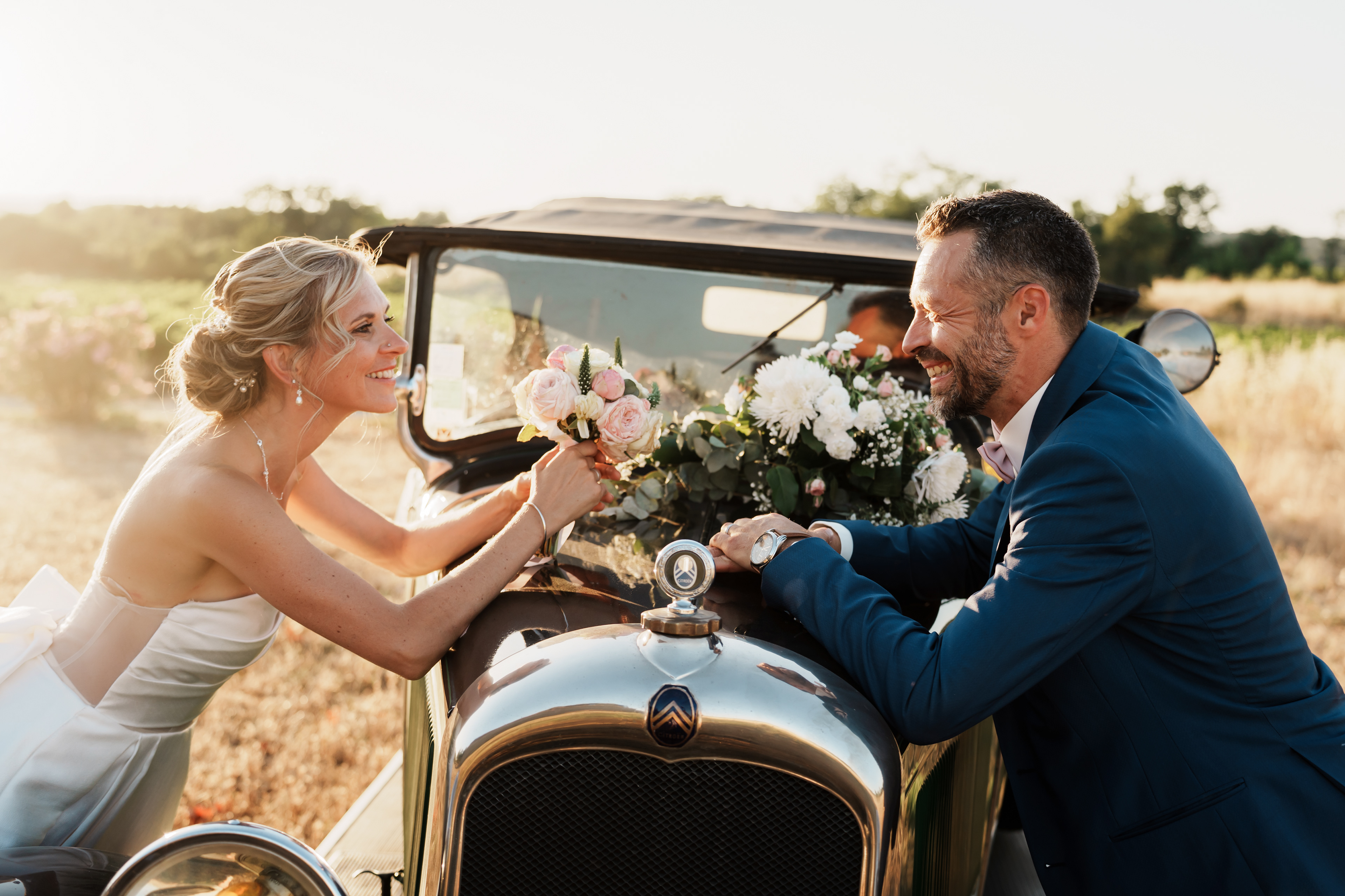 Les mariés se regardent face à face au-dessus du capot d’une voiture de collection au coucher de soleil lors d’une séance photo de mariage dans l’Hérault