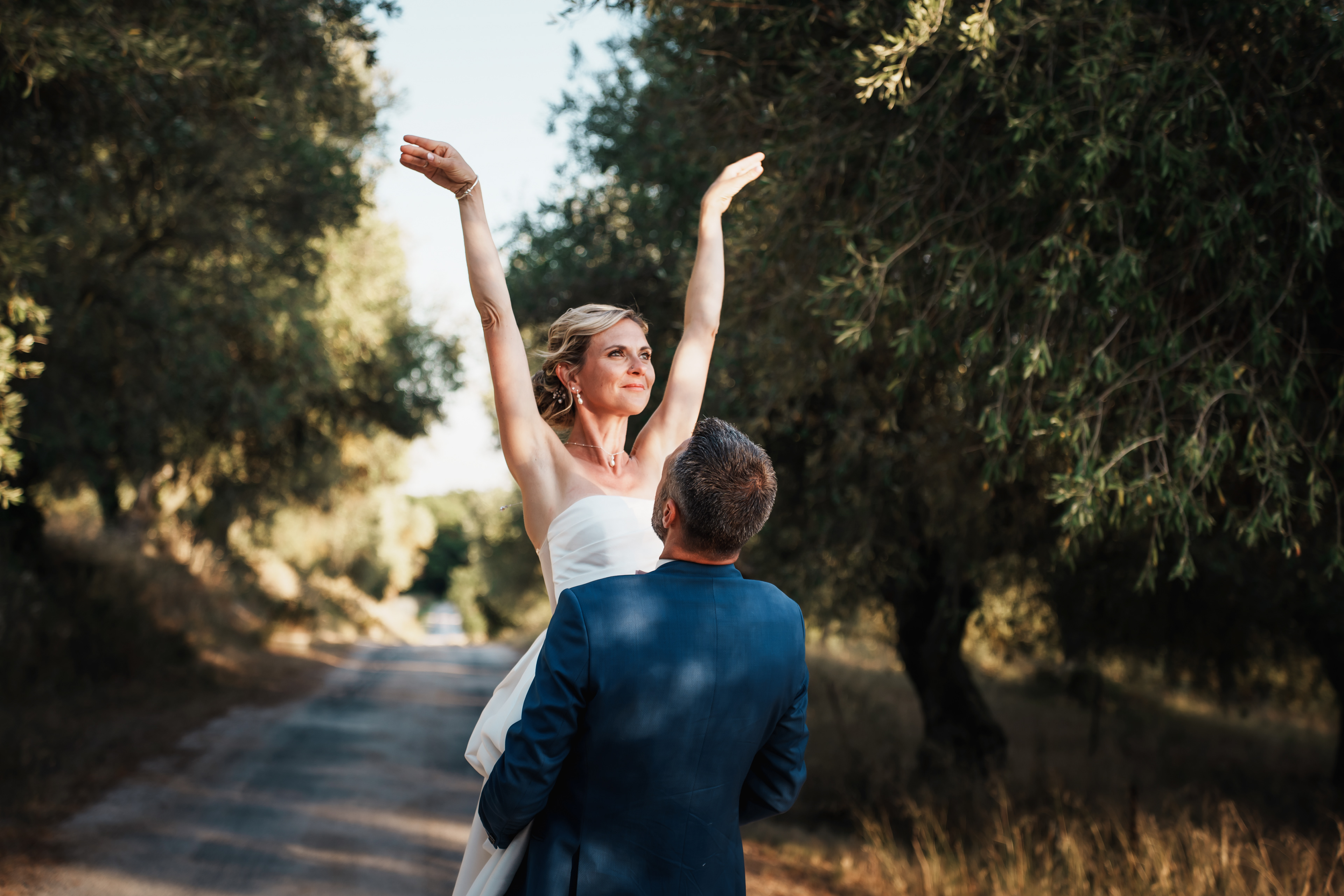 Les mariés dans une allée d’oliviers, le marié porte la mariée dans une scène pleine de complicité