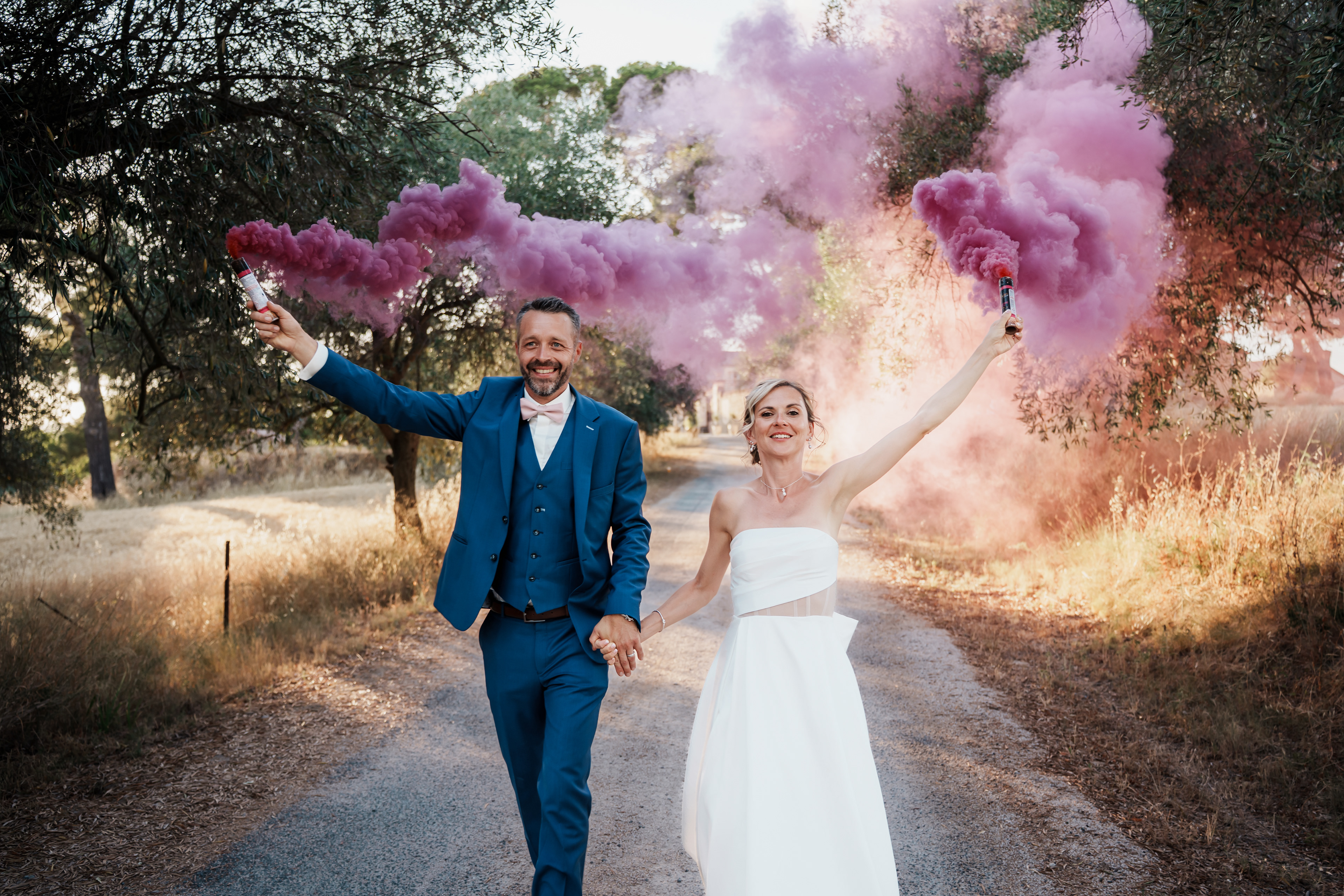 Couple de mariés avançant dans une allée d’oliviers entourés de fumigènes roses pendant la séance photo