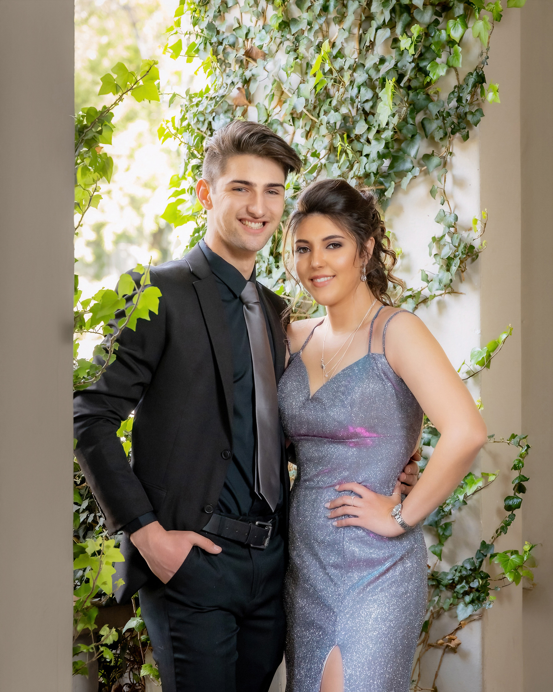 Ivan Muller Photography: A stylish matric farewell couple smiling, the girl in a sparkling silver dress with a high slit, and the boy in a black suit, standing by green ivy.