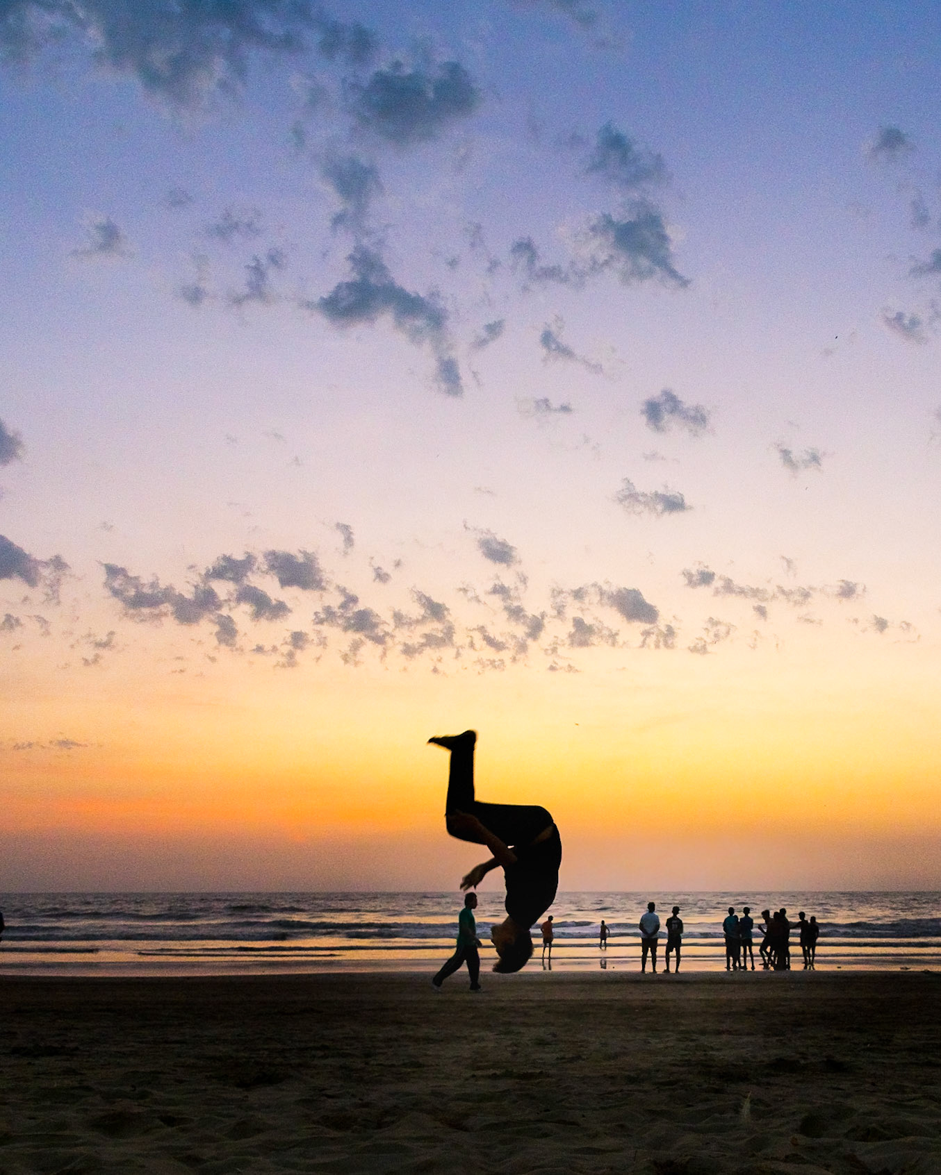 MUMBAI, INDIA – March 14 2019: On a hot summer evening youth performs somersaults, flips and handstands at the beach. Silhouetted at dusk with clouds in the sky.