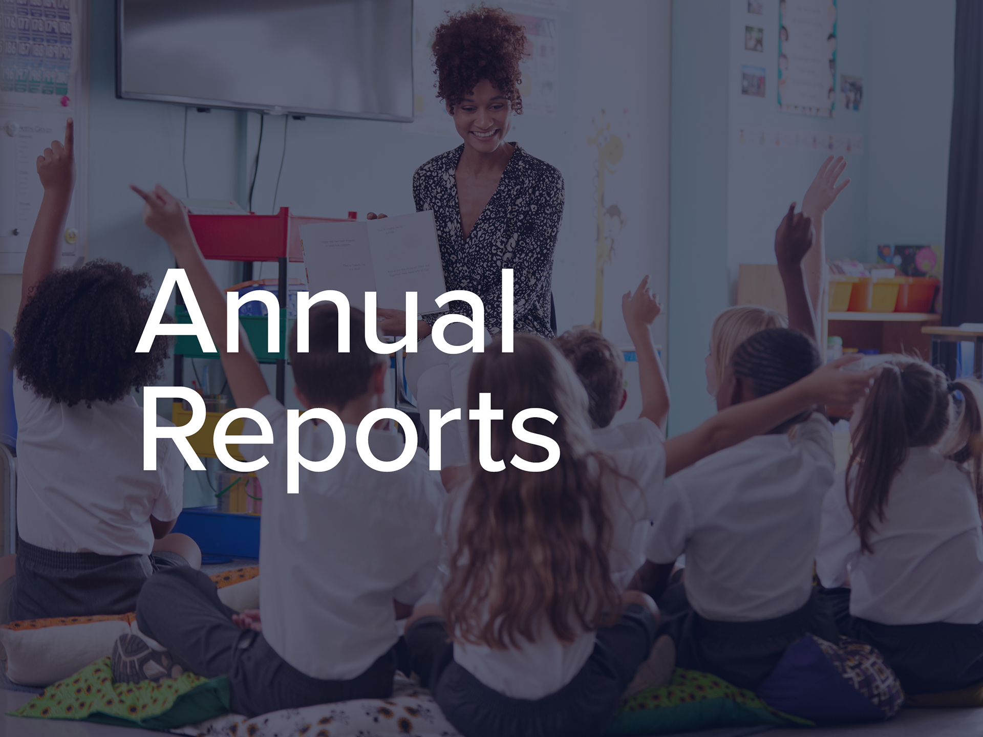 Annual Reports heading overlaid on a photo of a teacher with her students sitting in front of her with their hands raised to answer a question