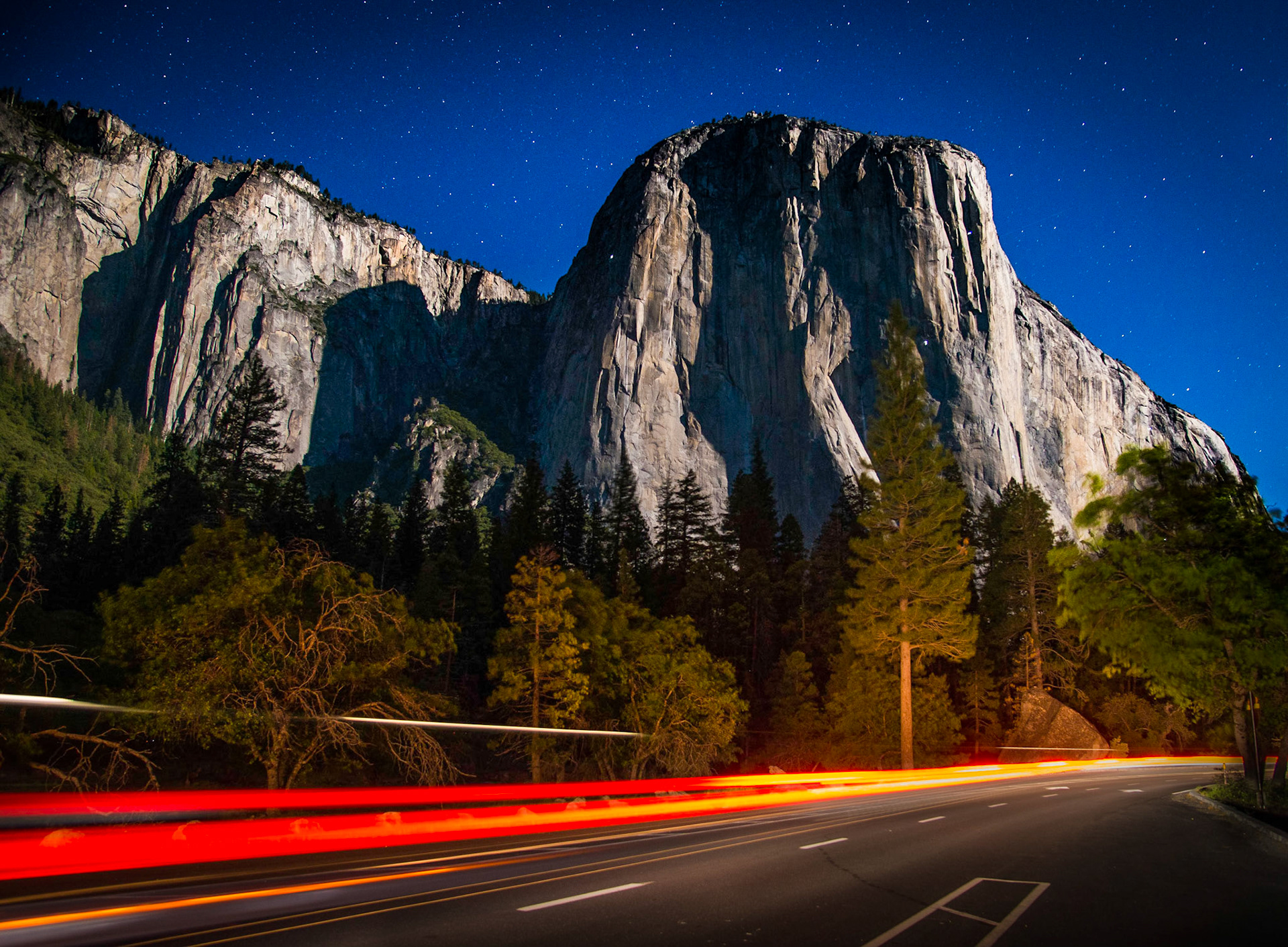 A long exposure captures the streaking tail lights of a car entering Yosemite Valley. Climbers' lights are visible on the face of El Capitan under a star filled night sky.