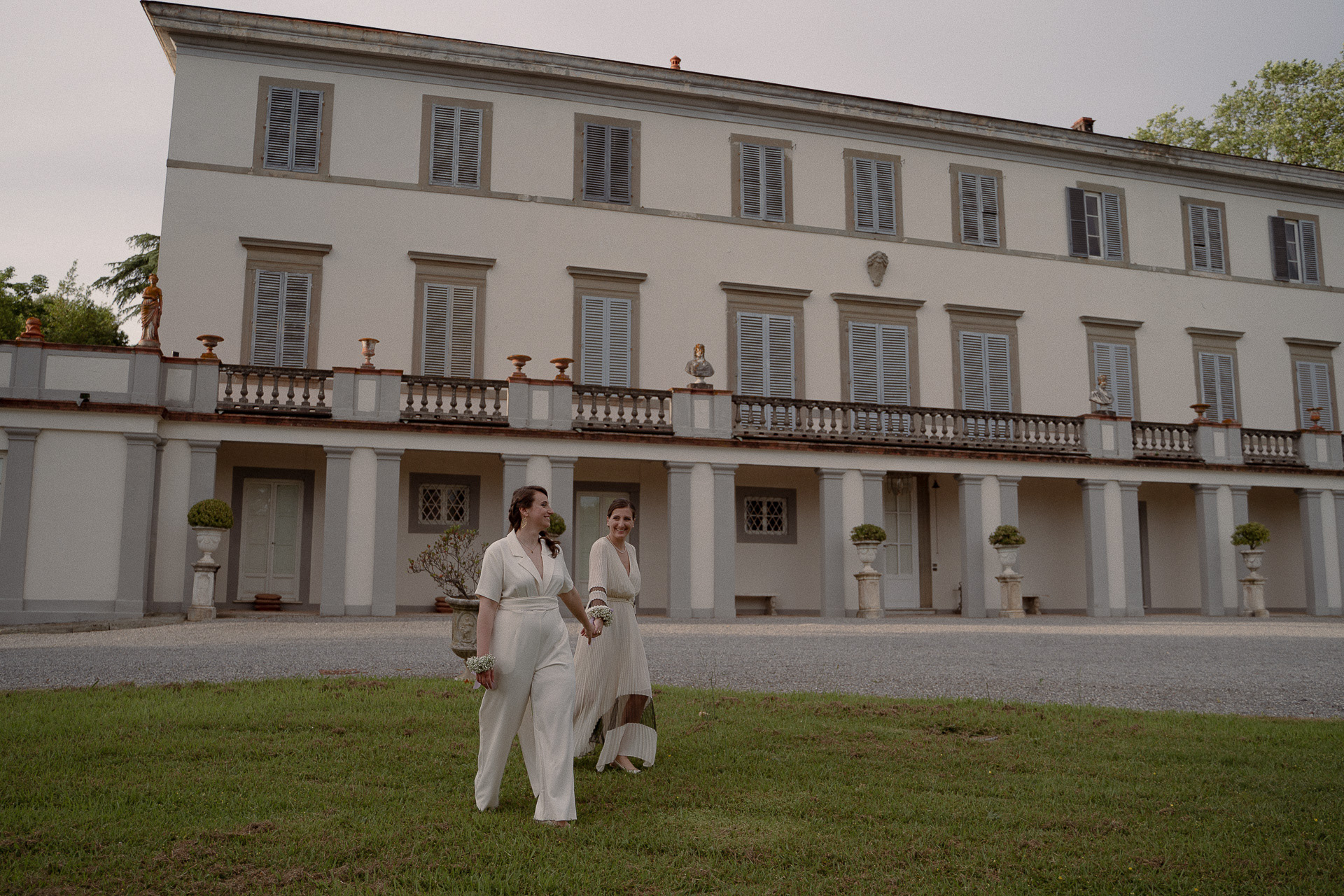 Fotografia di Arianna Macherelli, specializzata in Fotografia di Matrimonio, immagine orizzontale di due spose che camminano sorridenti mano nella mano con sfondo una villa