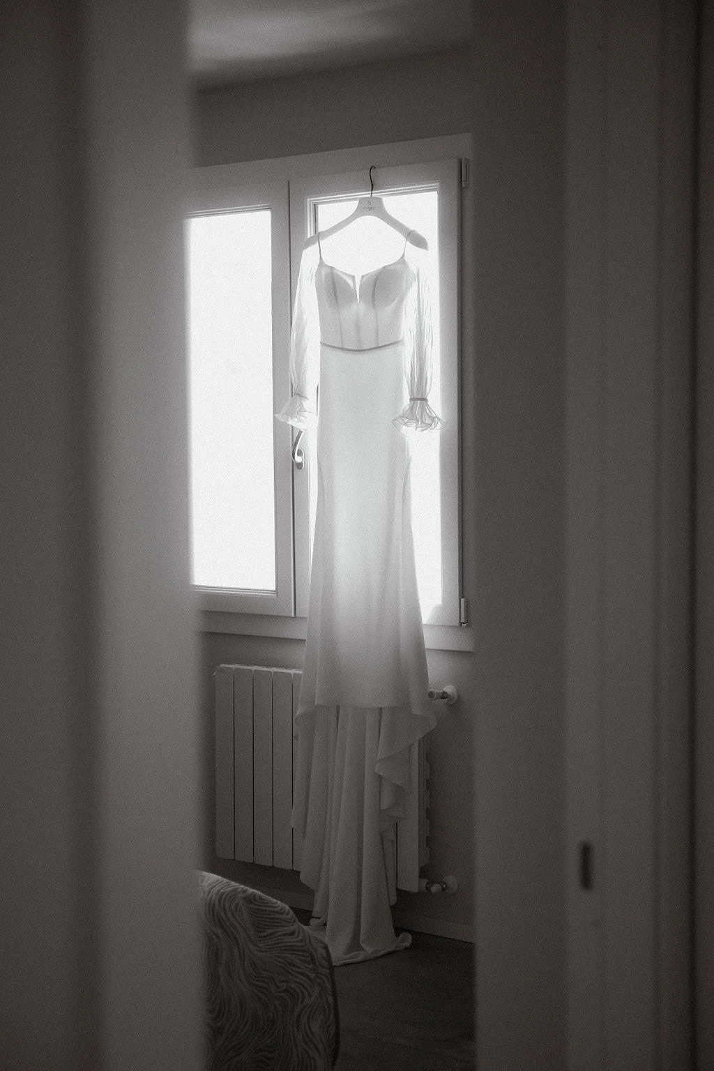 Fotografia di Arianna Macherelli, specializzata in Fotografia di Matrimonio, dettaglio in bianco e nero dell'abito da sposa appeso alla finestra in controluce