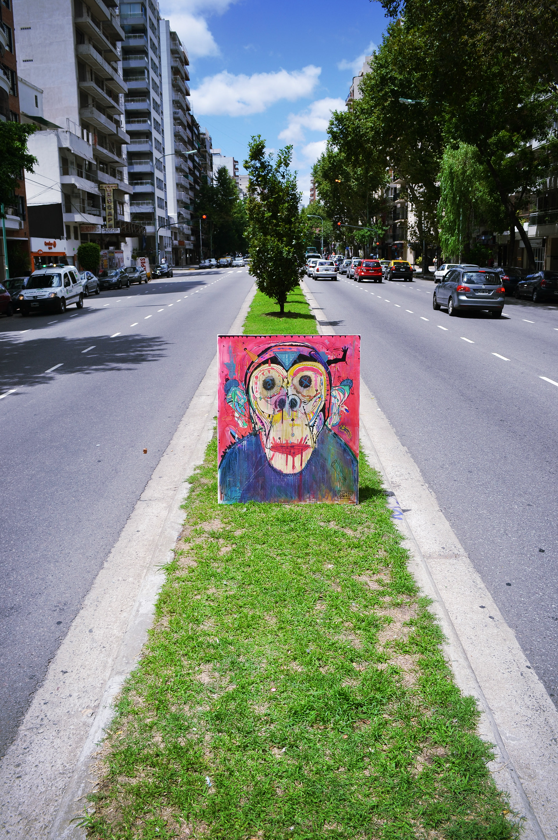 Axtor - Fotografía de una pintura expresionista de un chimpancé multicolor, colocada sobre el cantero central de una avenida arbolada.