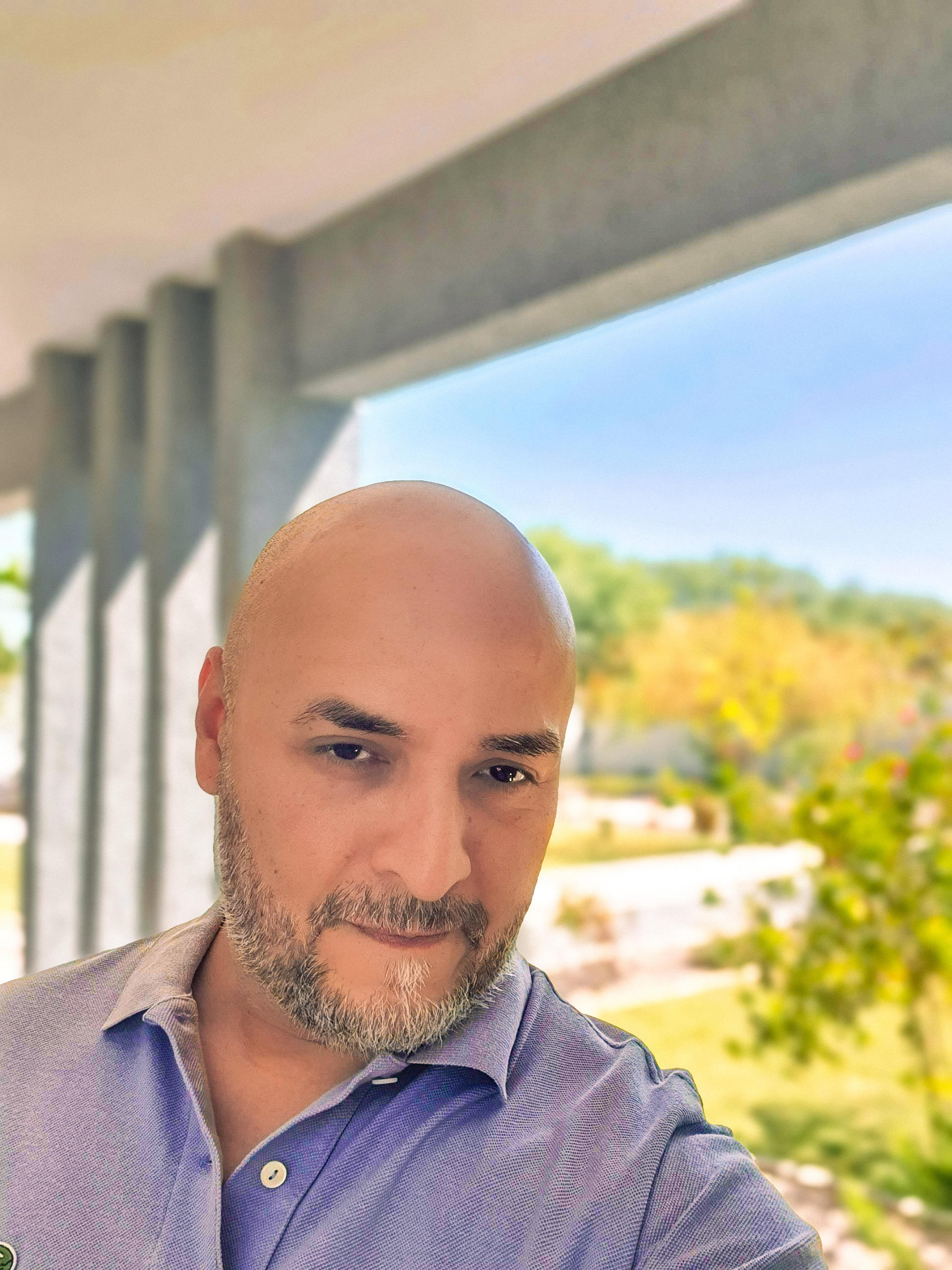 La imagen muestra a una persona calva con barba corta y canosa, vistiendo una remera de pique de color celeste claro con botones. El fondo incluye parte de una galería o porche con columnas de cemento y un paisaje exterior verde, con árboles y vegetación difuminados, dando la impresión de estar en un jardín bien cuidado bajo una luz natural intensa. La imagen transmite un ambiente tranquilo y soleado, con foco en Alberto Carrera que posa en primer plano