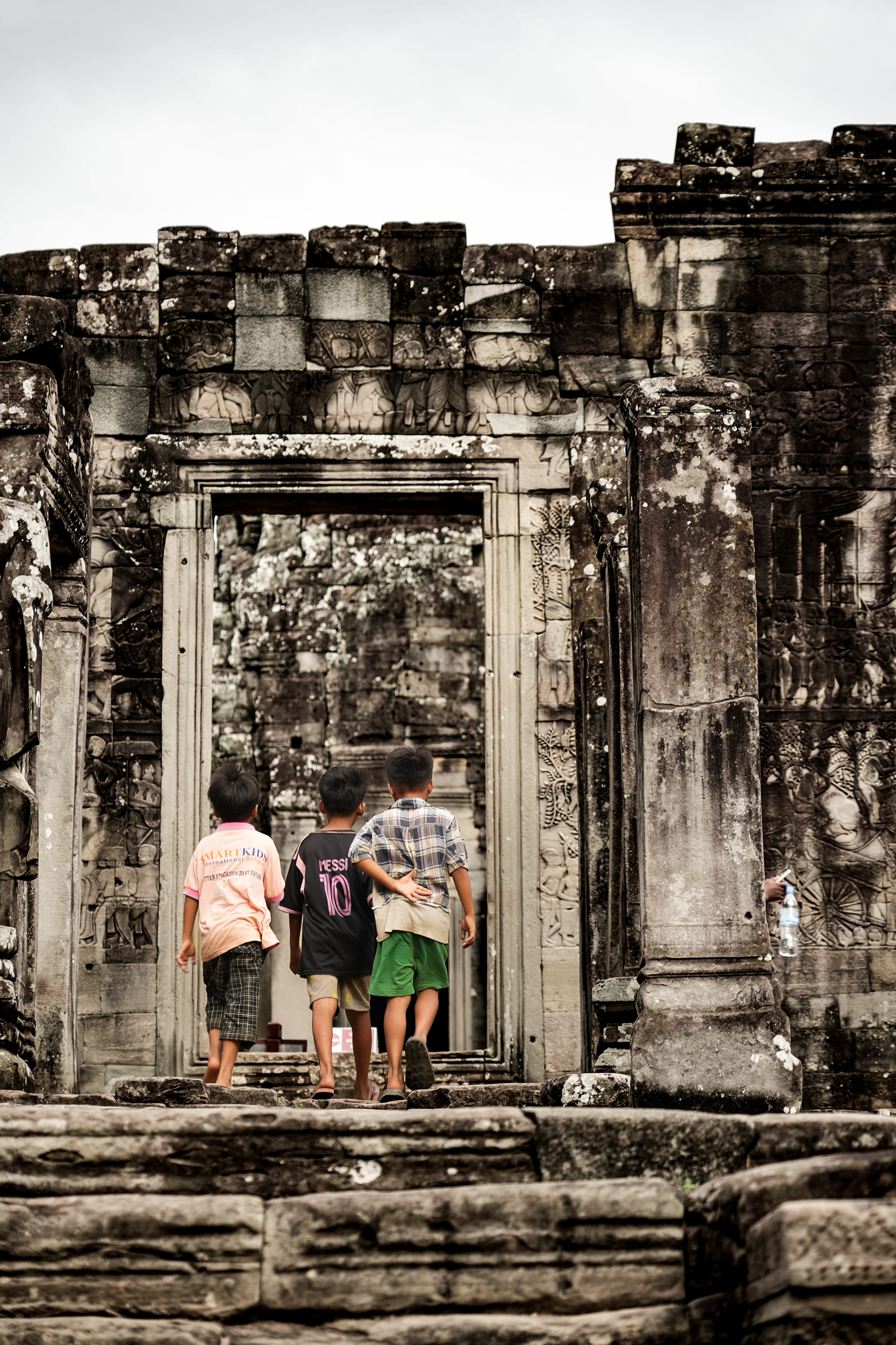 As I entered Bayon Temple, I came across Three Little Boys. They were running around, having fun, being kids. I glanced in their direction and saw a religious ceremony happening across the street. How interesting, I knew I was visiting temples, but little did I know that these temples, or their surroundings, still had a religious use. I assumed that these kids were waiting for their parents, too young to enjoy a religious ritual, preferring instead to play around with their friends. I wondered where the true privileges were: me, able to travel the world and visit some of its wonders, or the kids who knew this temple by heart and ran around on what seemed to be a usual Monday. Do the kids know on what grounds they are running? Where they are stepping? Did their parents teach them the importance of Angkor, the meaning of these temples? The faces looking out to the four cardinal points, watching over them like guardian angels. Is this why their parents are comfortable leaving them here? Or are the kids simply escaping what seems to them a boring religious ceremony? I wish I had asked them. But would they have answered if I had? We don't speak the same language. I wasn't able to ask them: What are your names ?