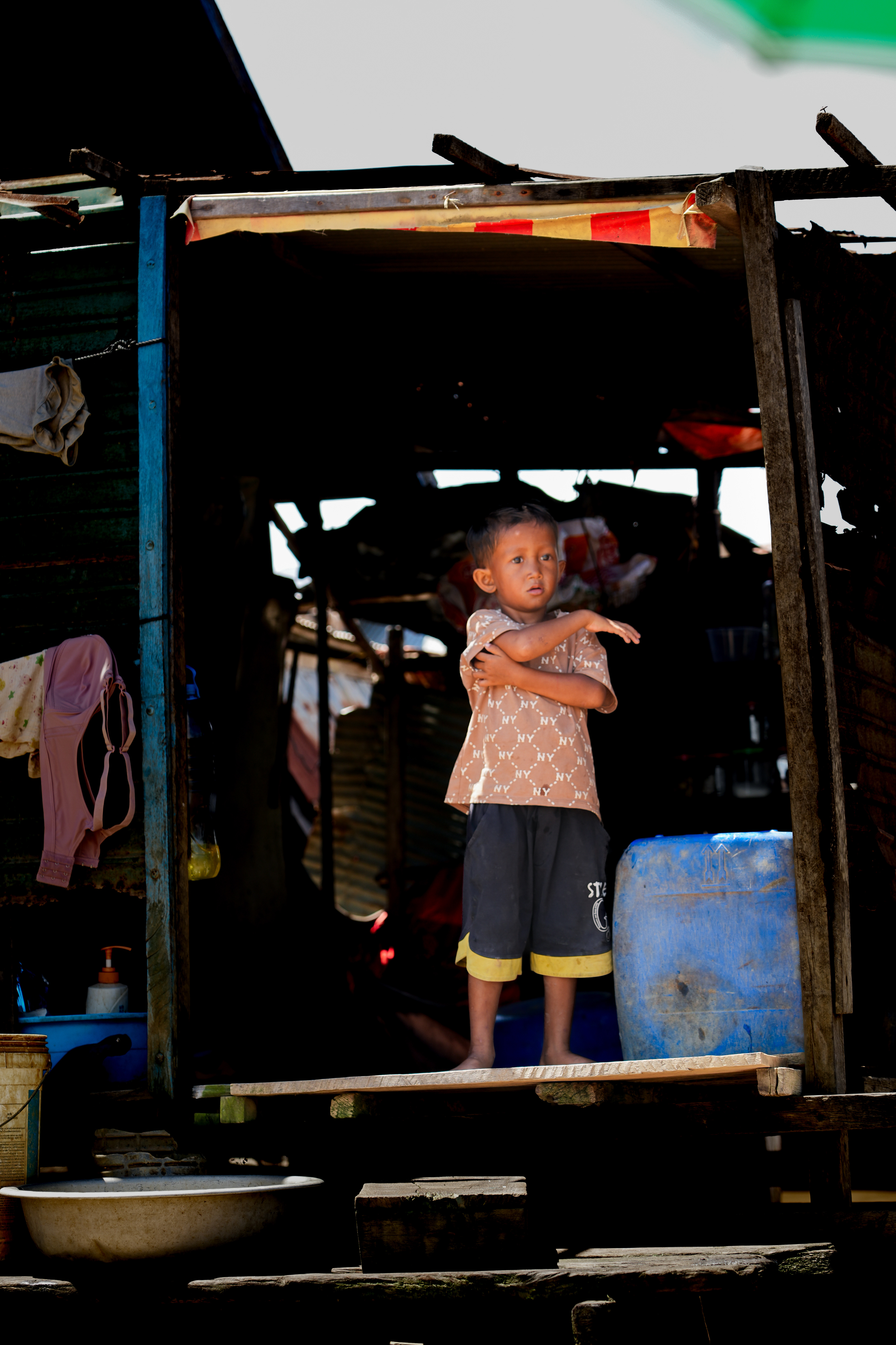 Cambodia. Chong Kneas Floating Village. Watching families in their home from a little boat on their river. I felt as if I was betraying my own morals. I wondered where the line was between intruder and visitor, between voyeur and photographer. I was there sitting on a barque covering myself from the heat with an umbrella, while they remained in their stilted homes above the water, waiting to see if dinner will be served. It felt wrong watching them, I saw this Little Boy looking confused, I don't know what he's waiting for. Is he waiting for his parents to return, or for his next meal? Does he eat enough at night? Does he notice me, sitting in a boat, photographing silently from the water? When does he go to school, or does he even go at all, with the nearest village a long way off? What grade is he in? How old is he? And what is his name?